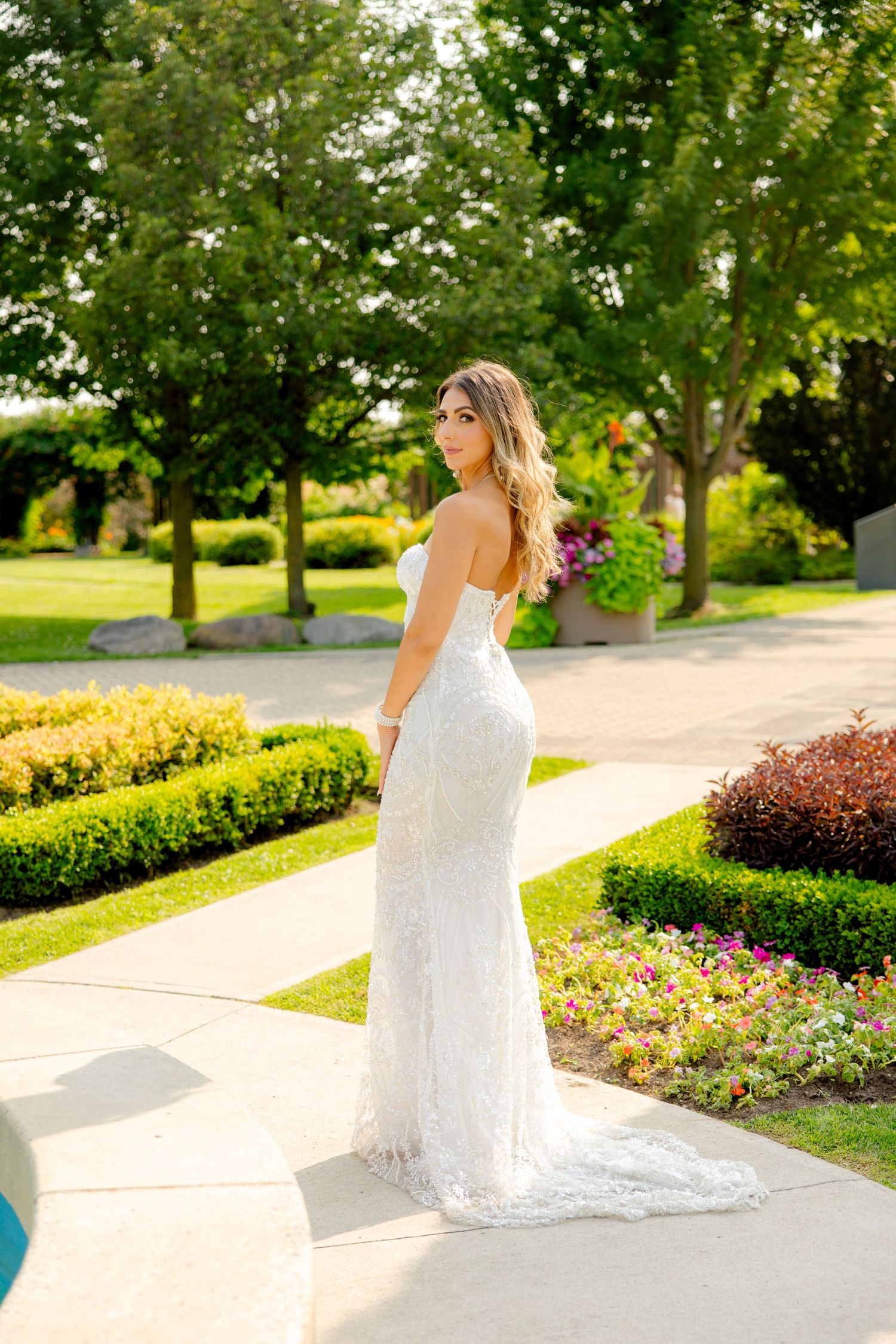Bride wearing form-fitting lace gown poses in landscaped garden with trailing train.