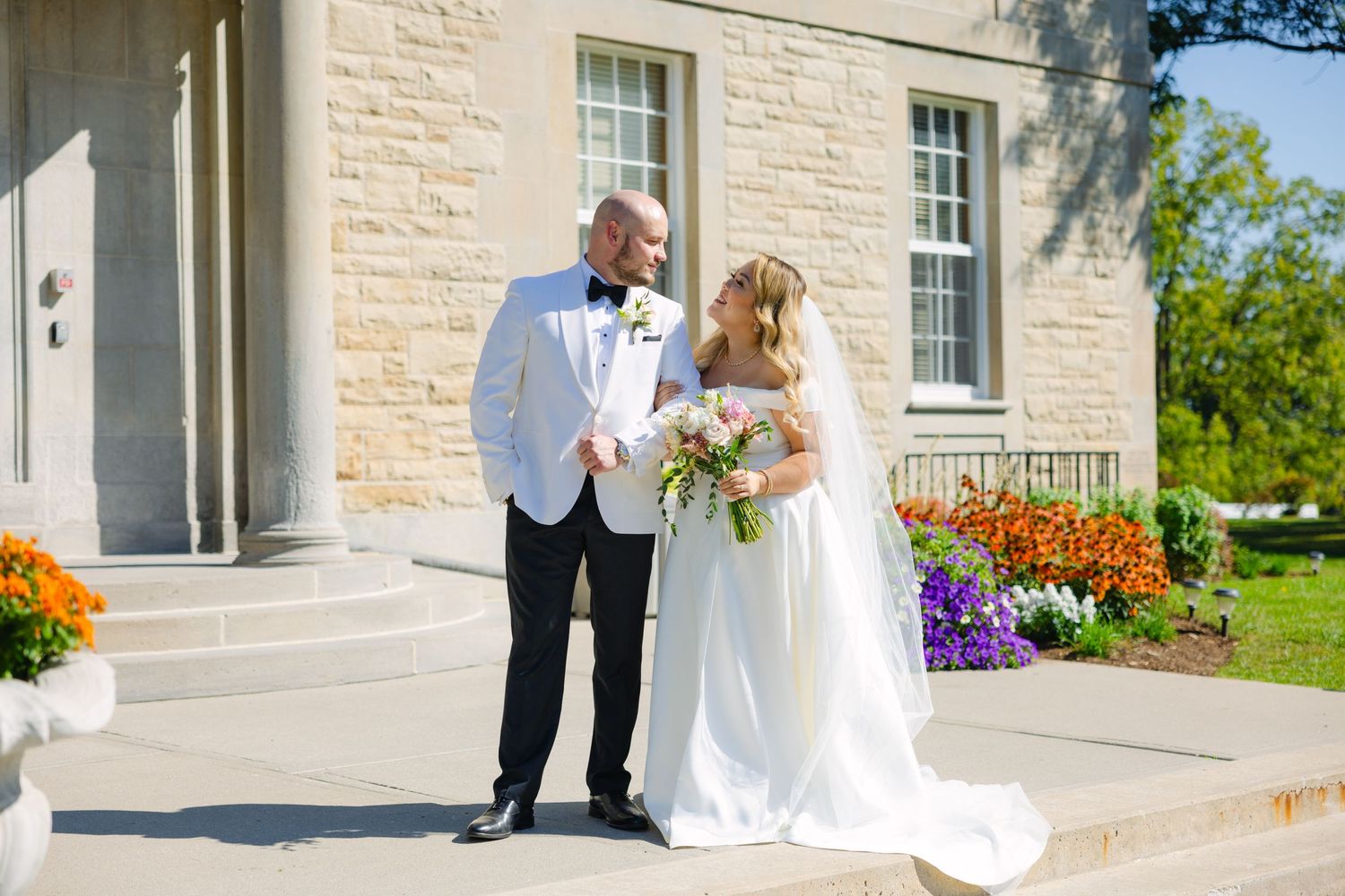 Sunny wedding portrait with couple standing by stone building and vibrant flower beds.