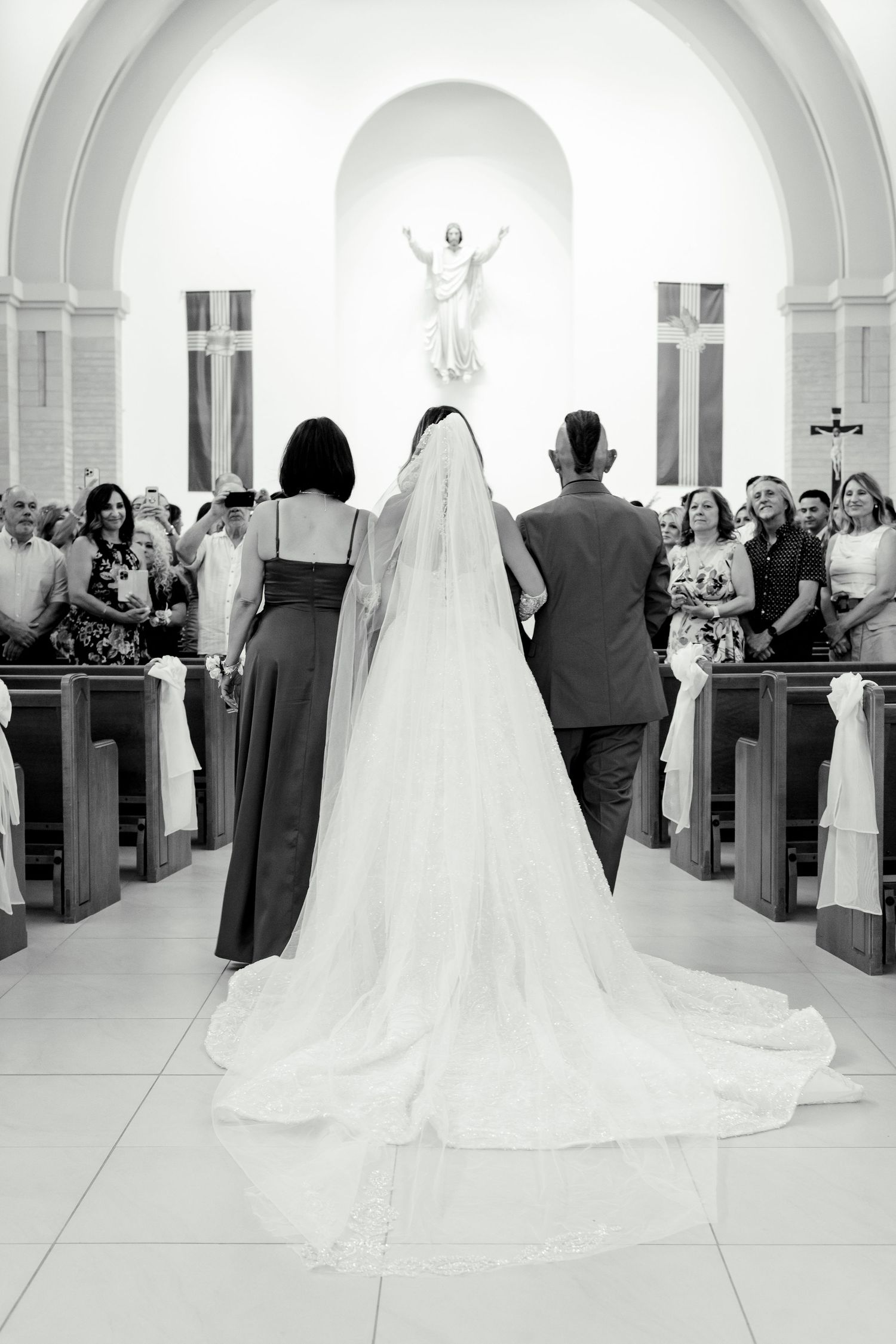 A black and white photo of a wedding processional in a church with a statue of Jesus at the altar.