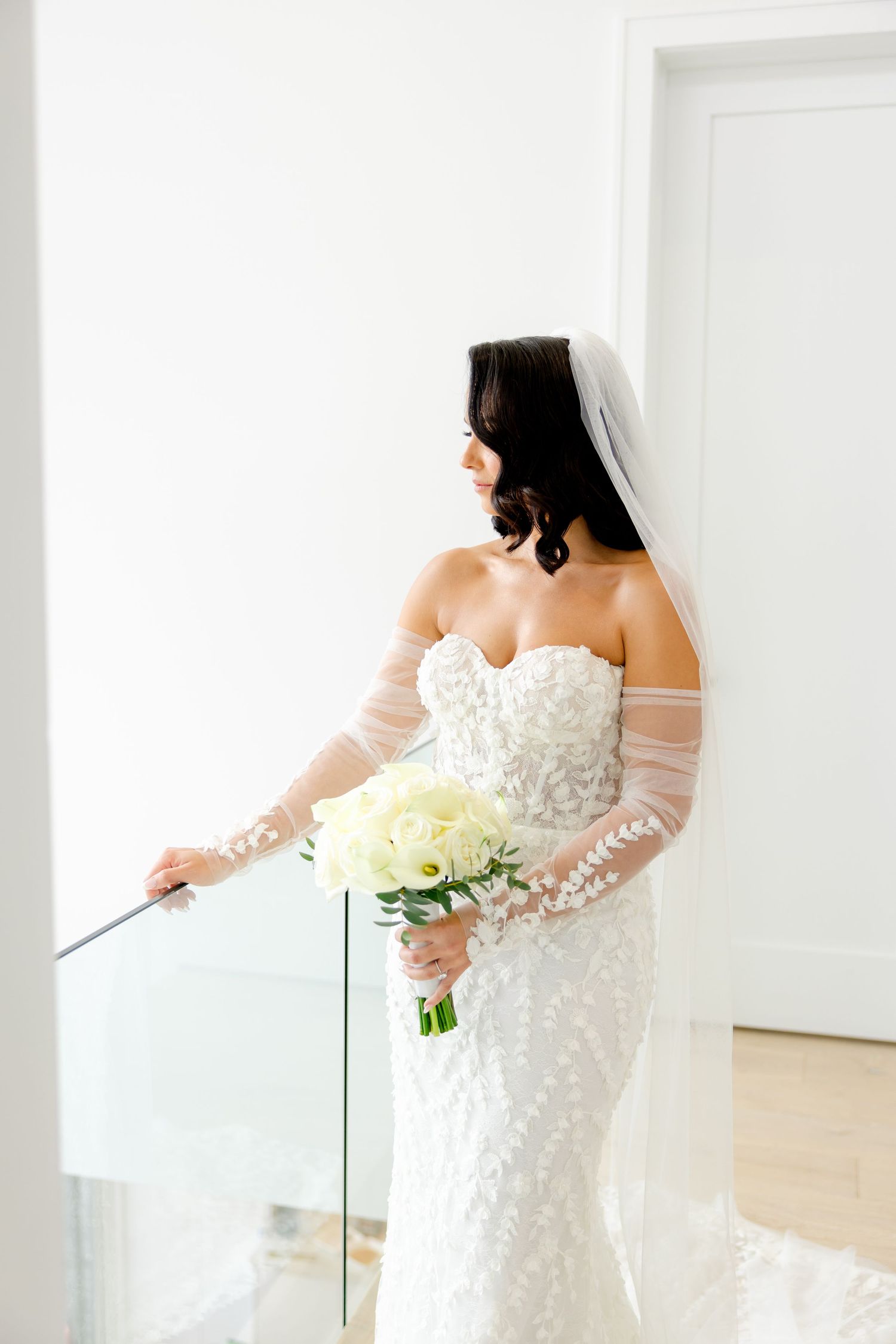 An elegant bridal portrait featuring an off-shoulder lace gown and white rose bouquet beside glass railing.