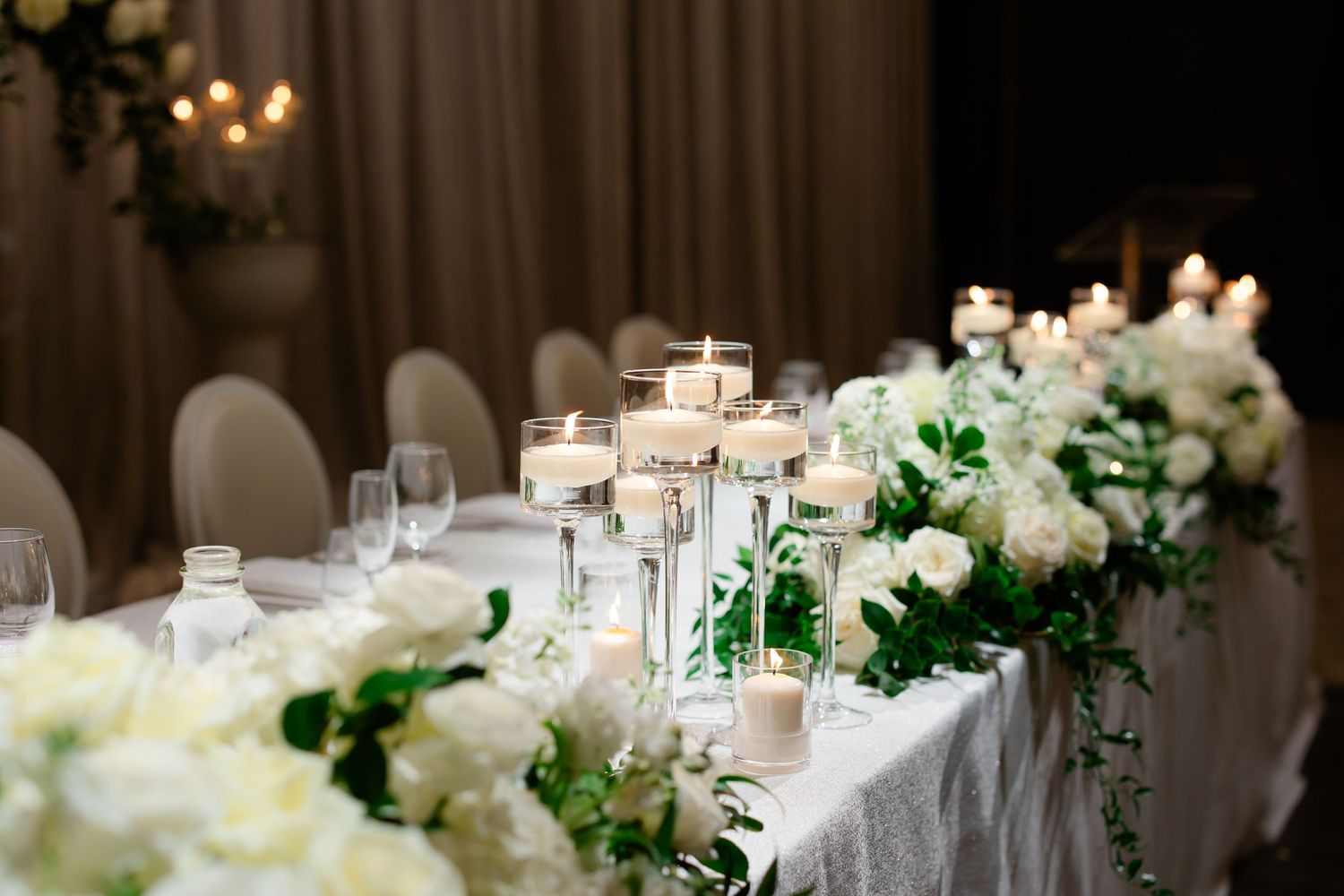 White floral arrangements and glowing candles line an elegantly set wedding reception table.