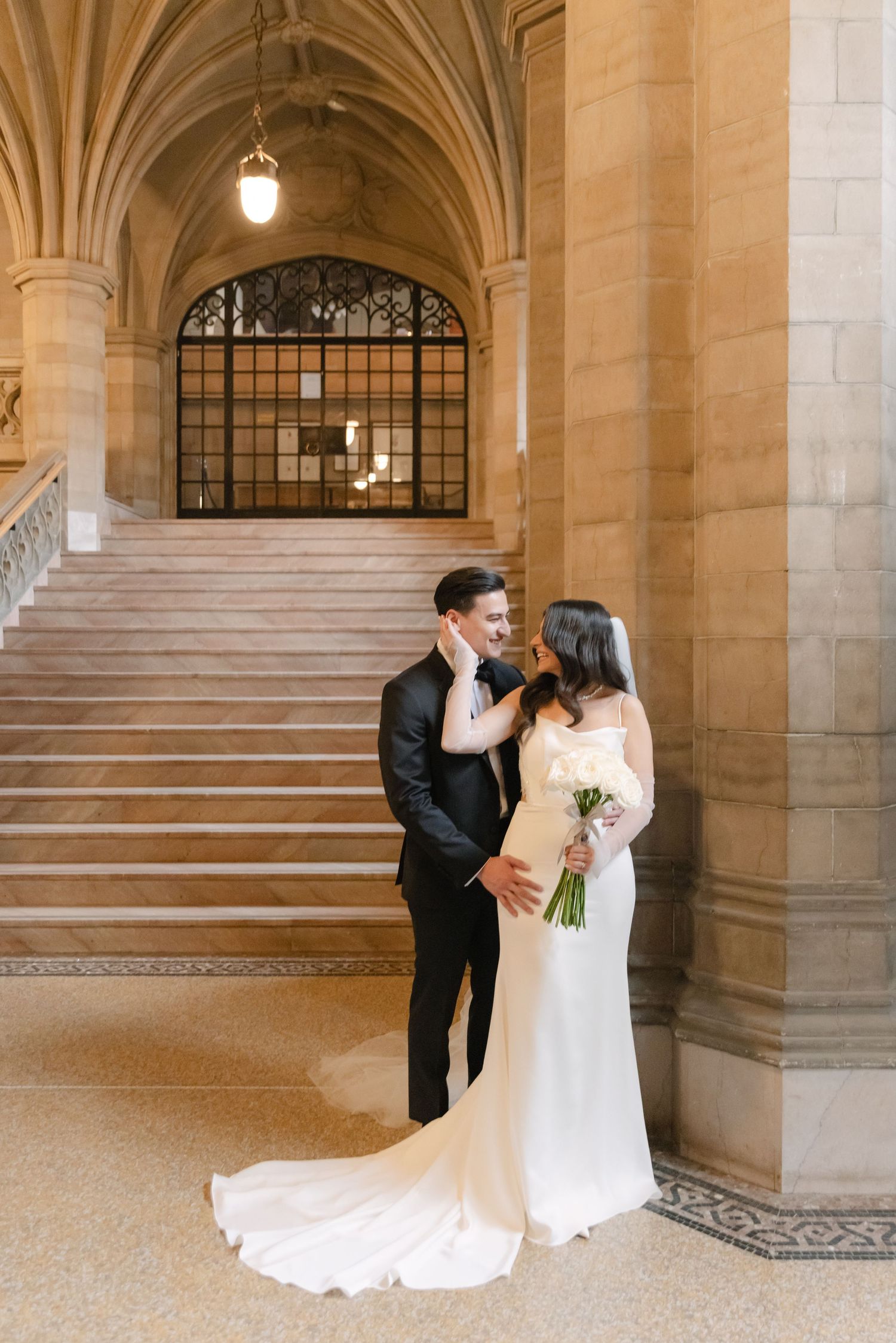 Romantic wedding photo of couple embracing by grand marble staircase with gothic architecture.