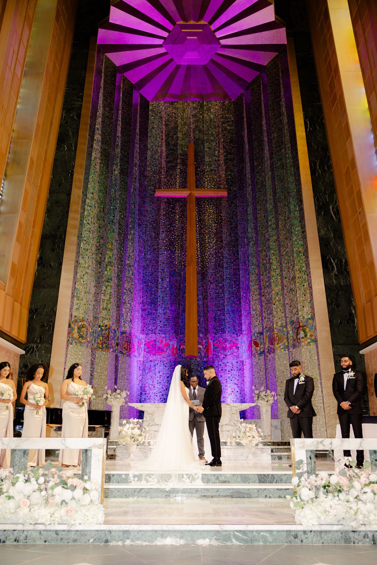 Wedding ceremony beneath dramatic purple-lit church ceiling with cross and floral arrangements.