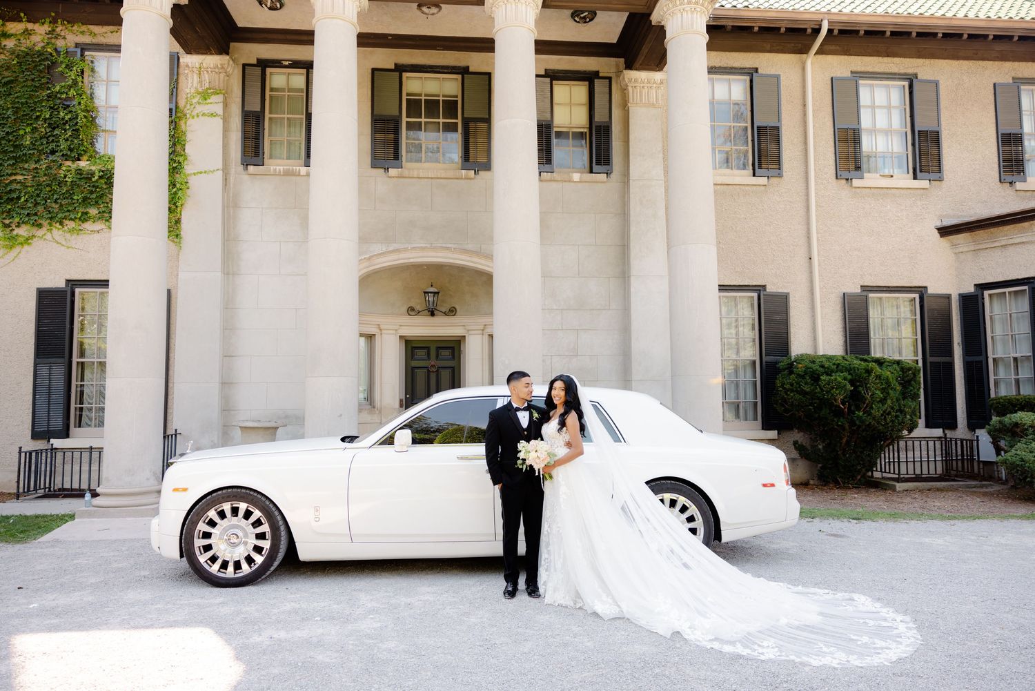 Wedding couple poses next to luxury white car in front of elegant mansion with white columns.