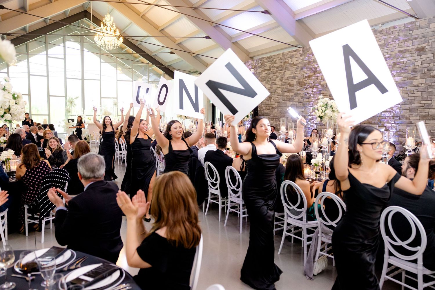 Wedding guests hold up letters spelling out a word during a celebratory reception in a modern venue with vaulted ceilings.