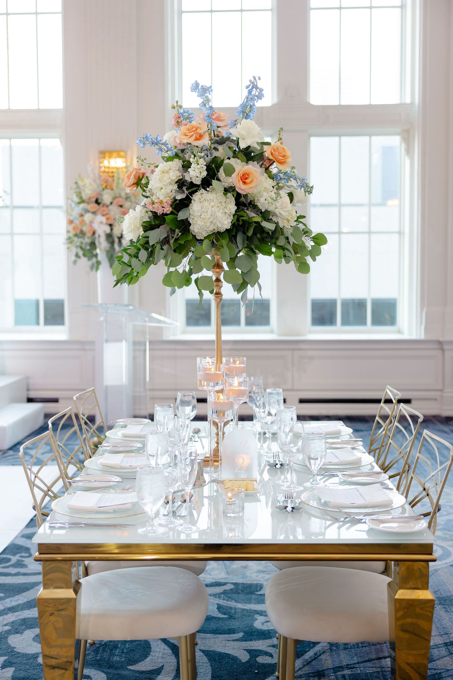 Wedding reception tables with gold accents, white linens, and lush floral centerpieces in a bright ballroom setting.
