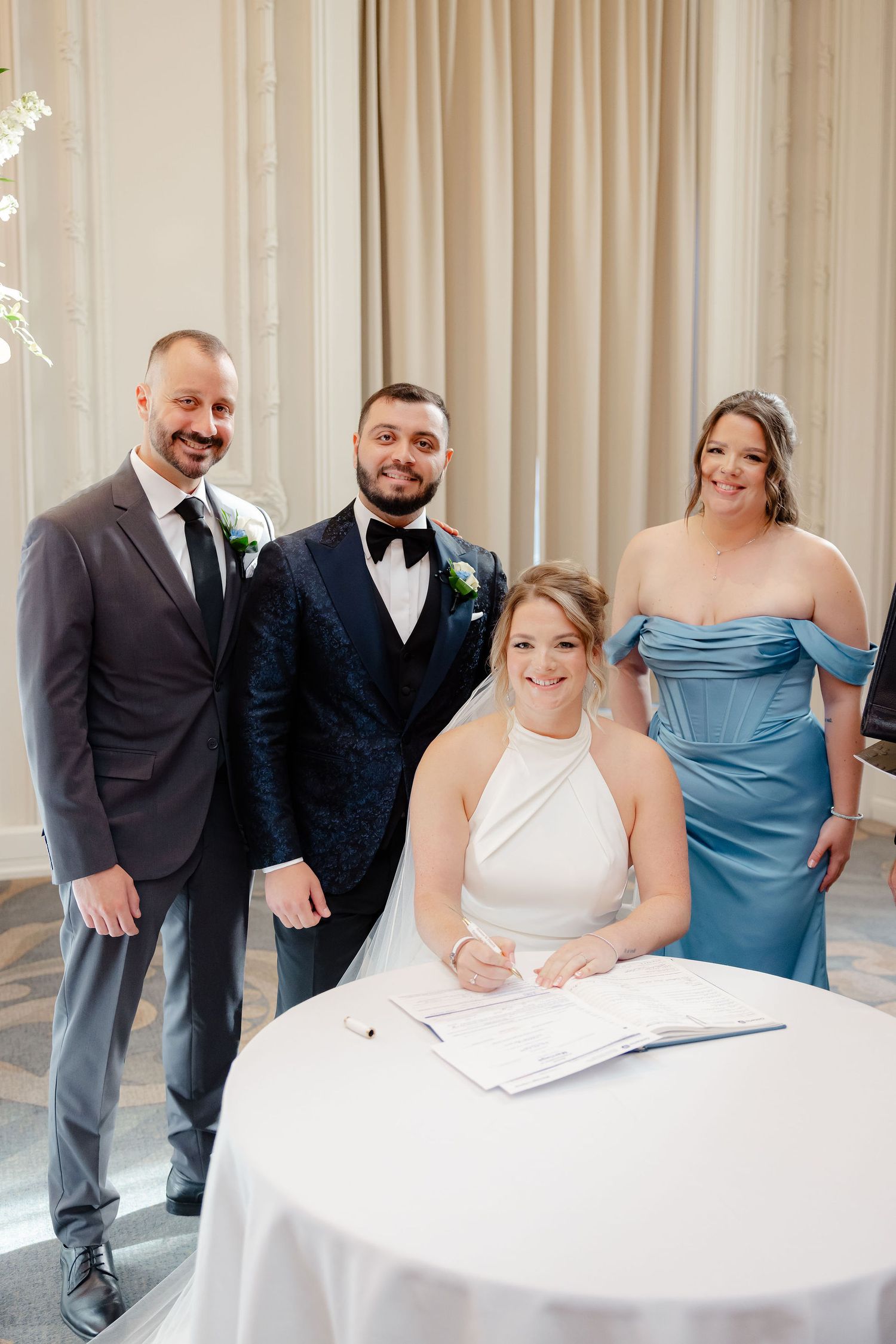 Wedding party gathers around a white table for document signing in an elegant venue.