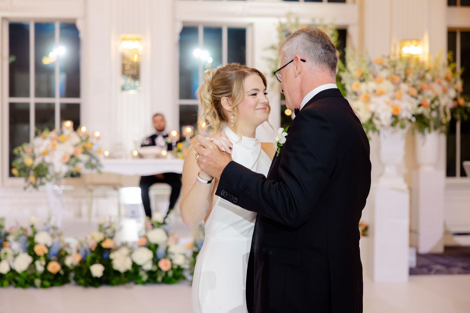 A couple shares an elegant dance moment at a wedding reception decorated with floral arrangements and soft lighting.