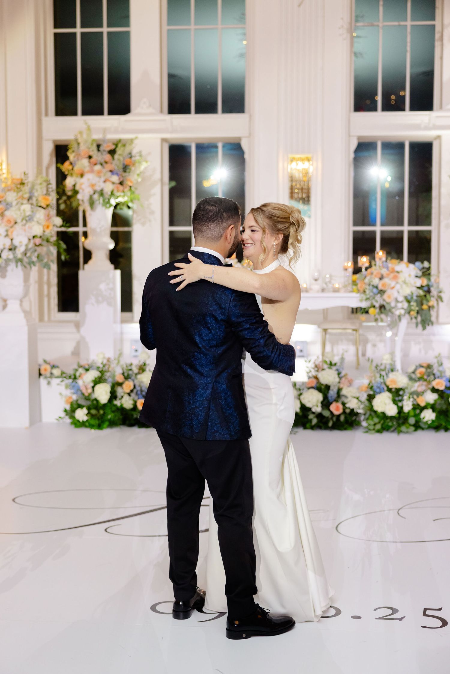 Newlyweds dancing together in an elegant white ballroom decorated with floral arrangements.