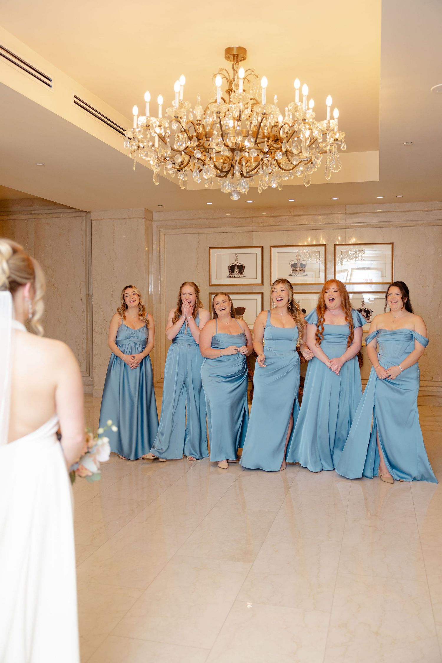 Bridesmaids in matching light blue dresses pose together in an elegant room with crystal chandeliers.