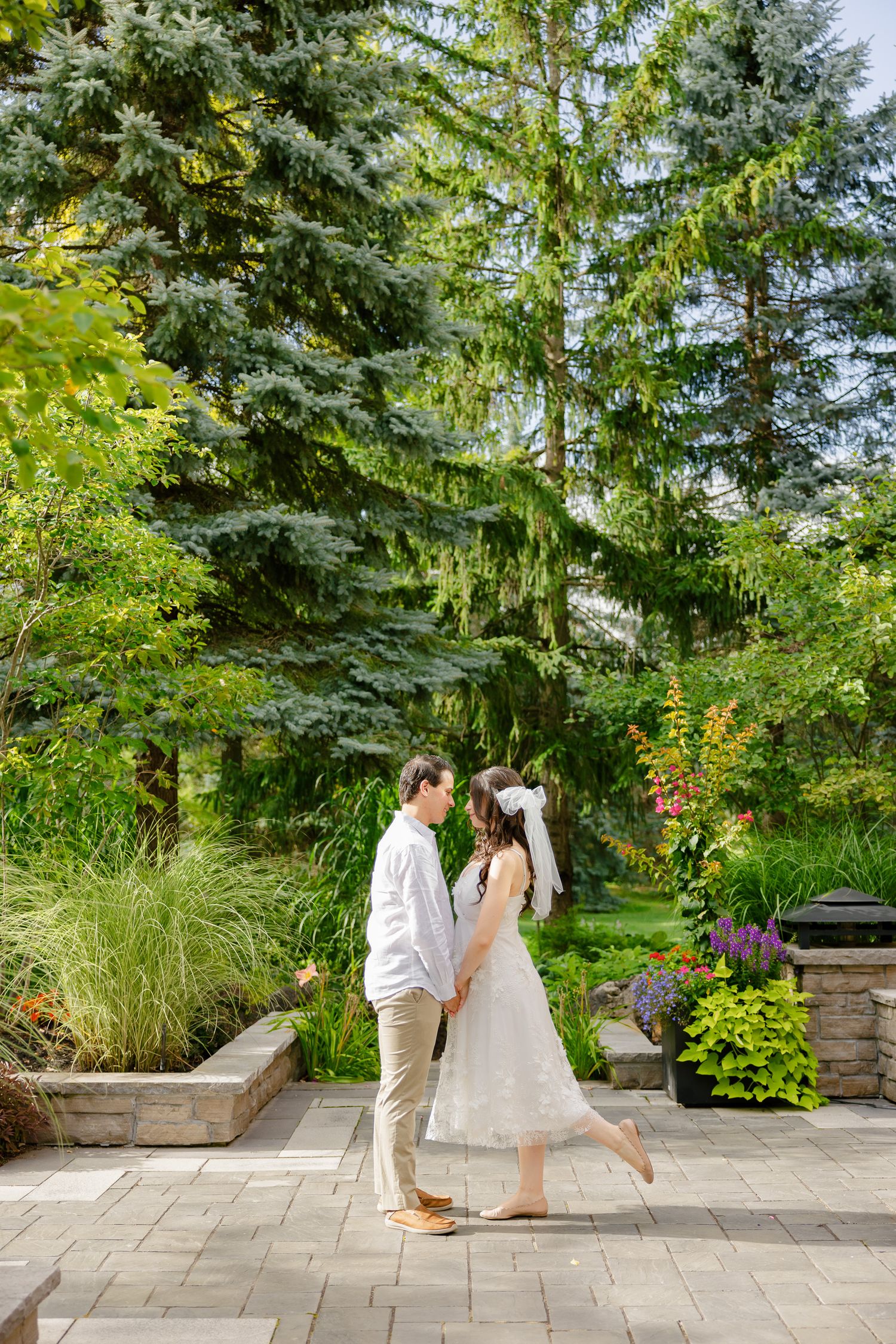 A romantic dance moment between a couple on a stone patio surrounded by evergreens and flowering plants.