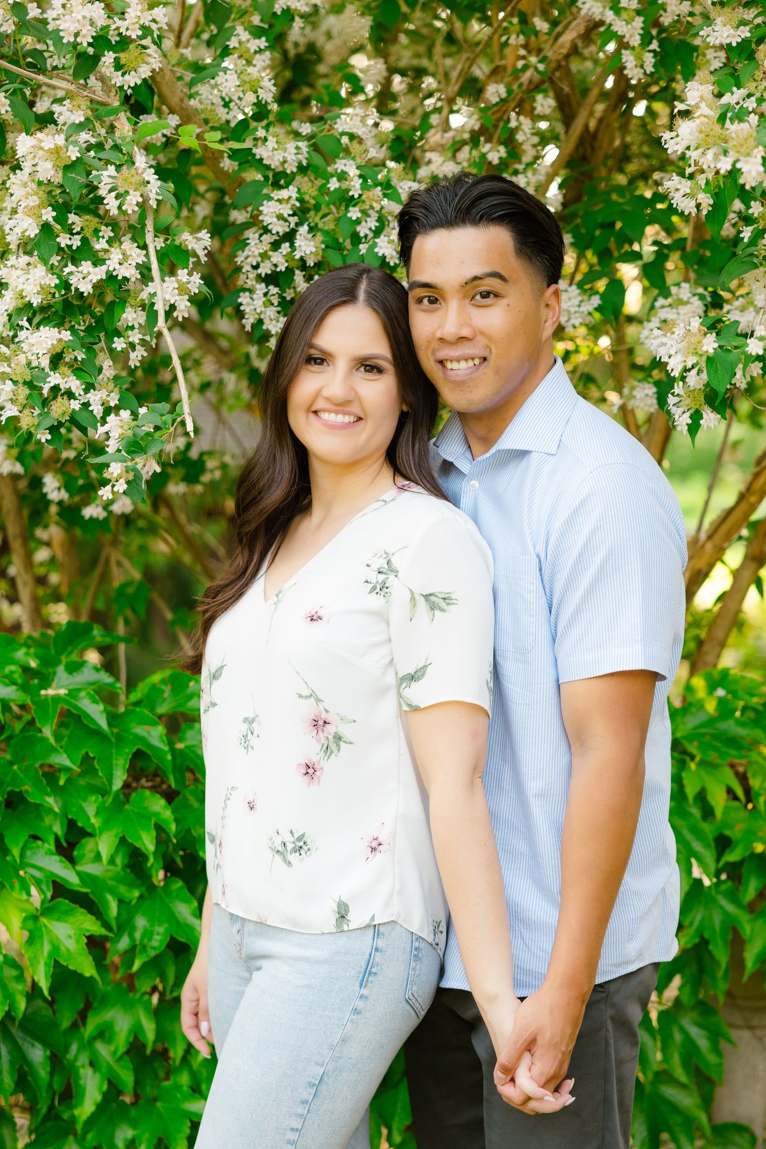 Couple holding hands and smiling while standing against flowering trees in spring garden setting.