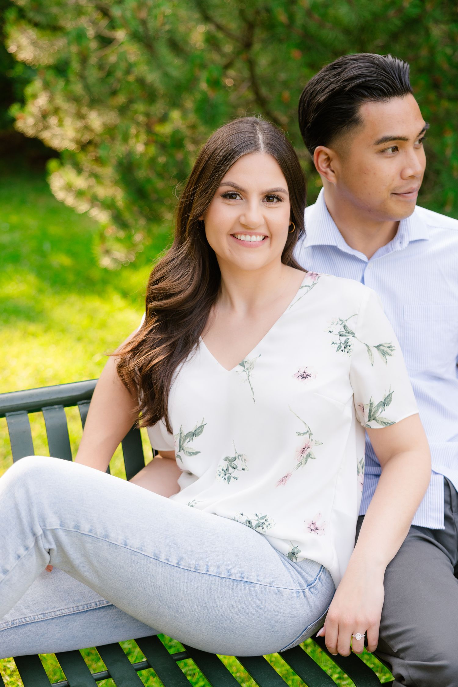 A couple poses together on a green park bench with sunlit foliage in the background.