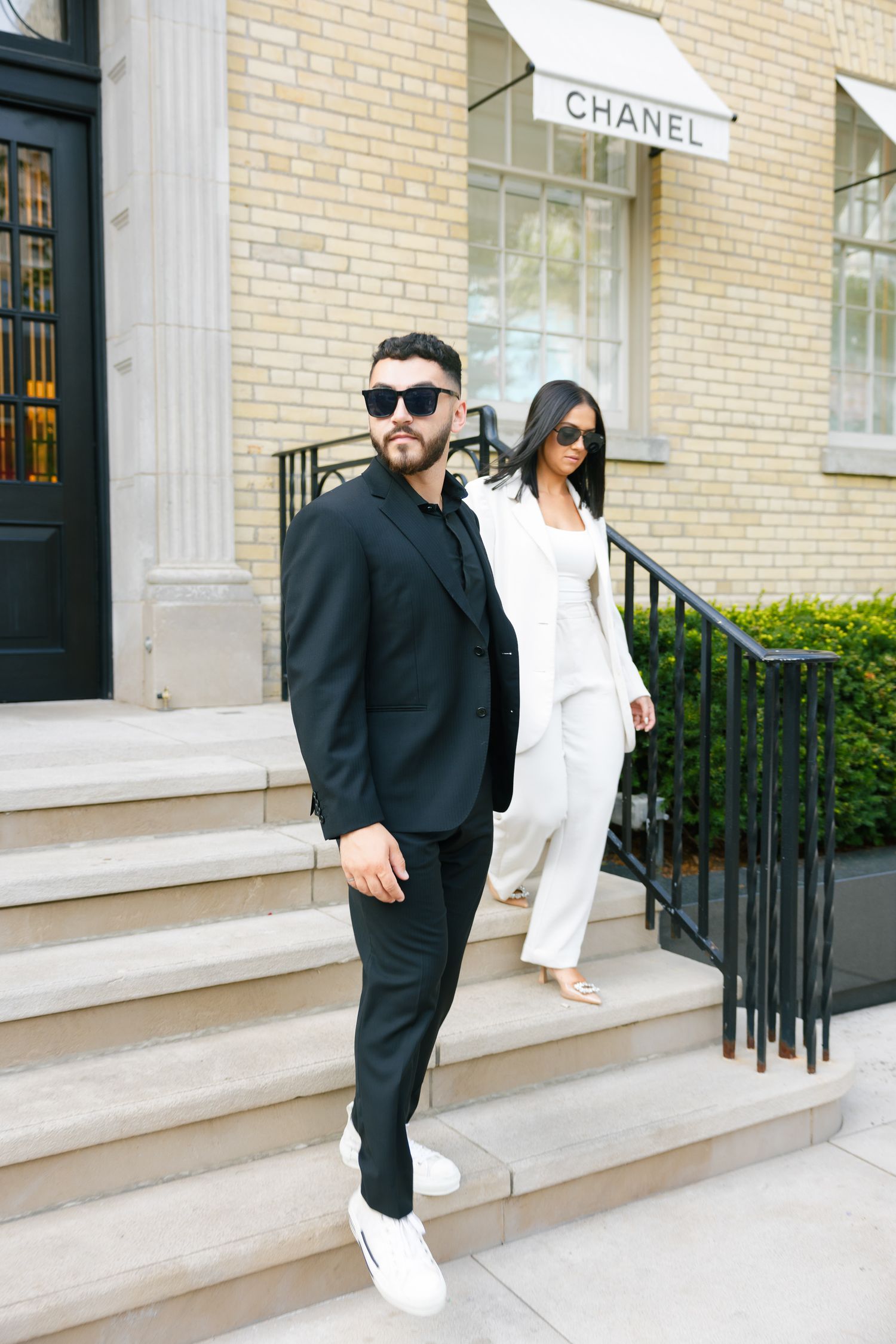 Fashion-forward pair in monochromatic outfits pose outside luxury retail store.