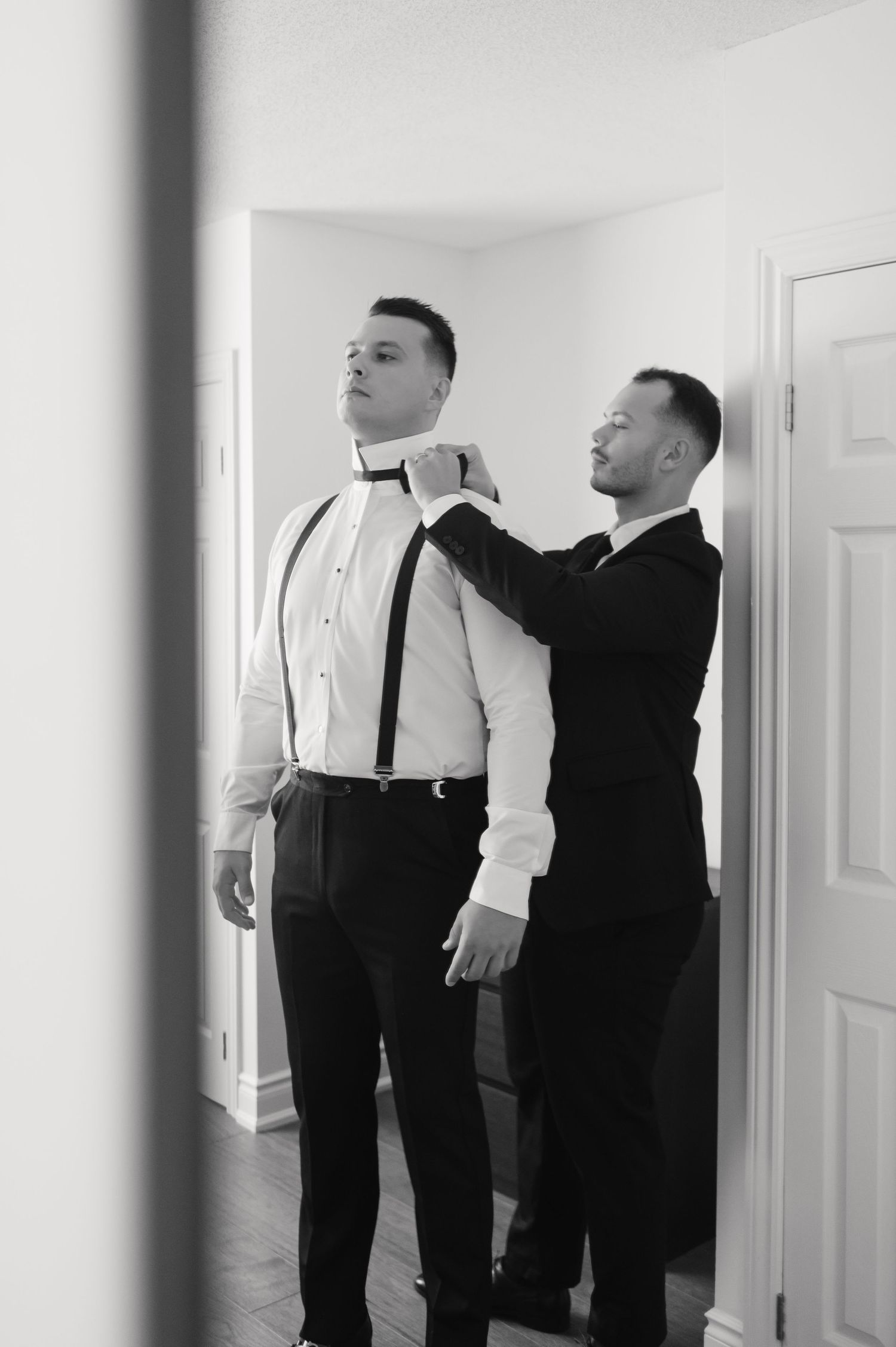 A formal preparation scene showing someone helping adjust a bow tie and suspenders.
