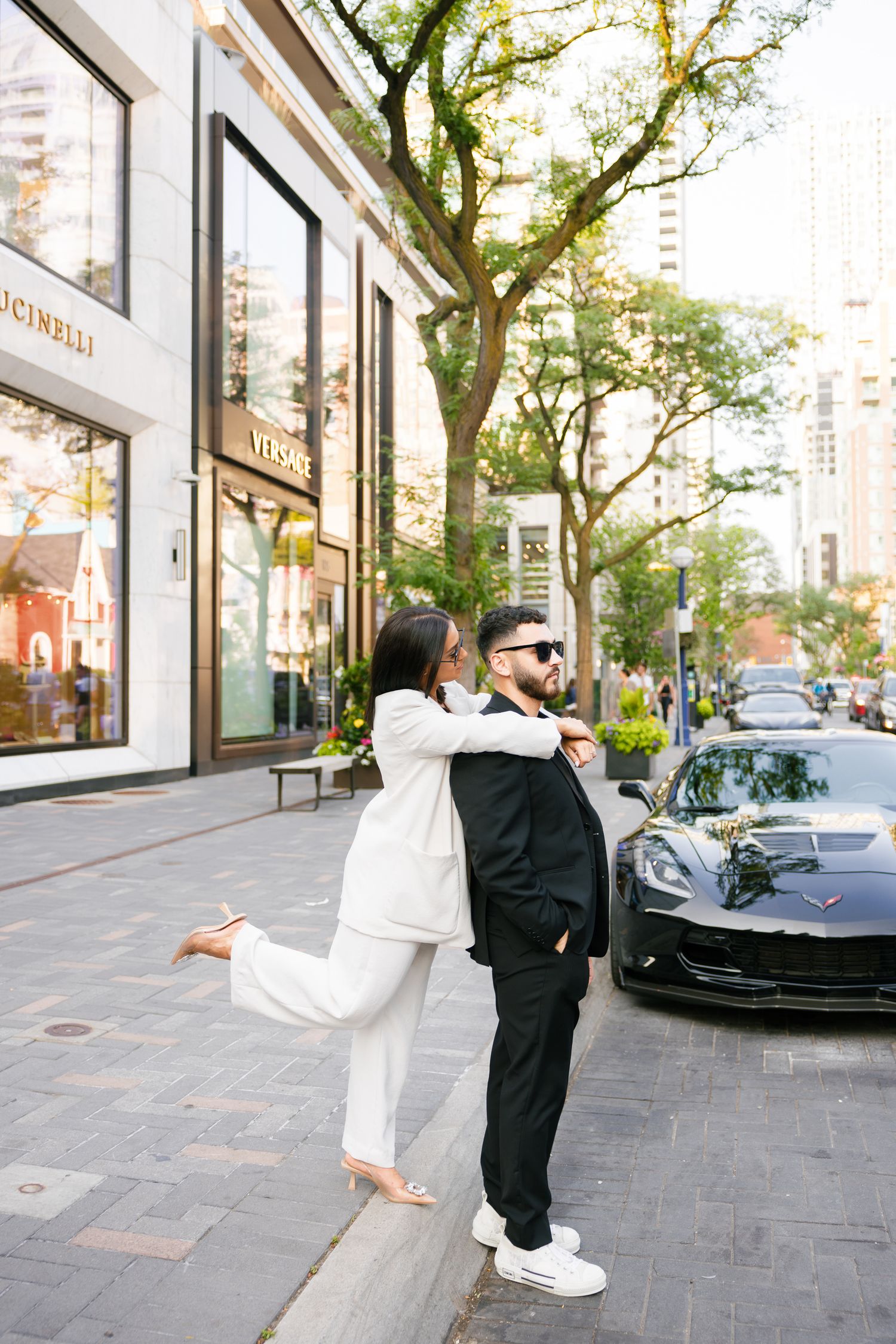 Playful street fashion photo with couple in contrasting suits near luxury car and storefronts.