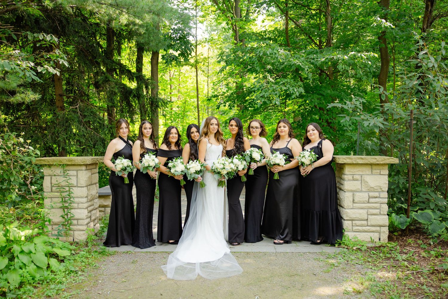 An elegant wedding group portrait with bridesmaids in black dresses standing between stone pillars.