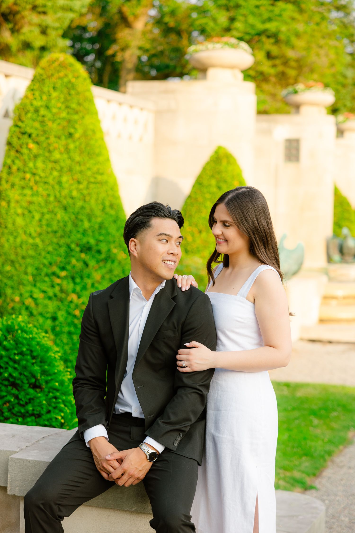 A couple sharing an intimate moment on a stone bench in a formal garden with topiary bushes.