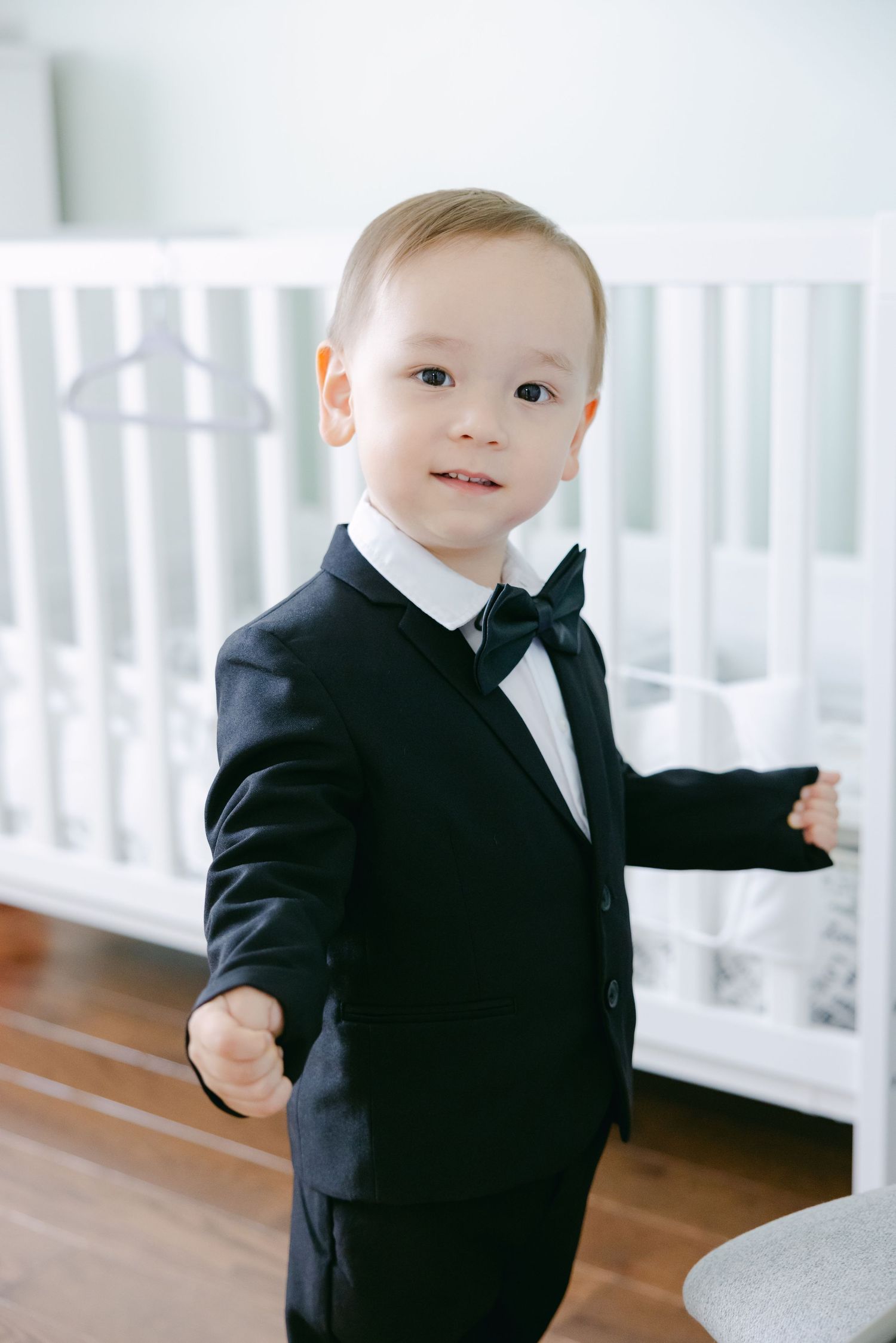 Toddler dressed in formal black tuxedo with bowtie standing by white crib.