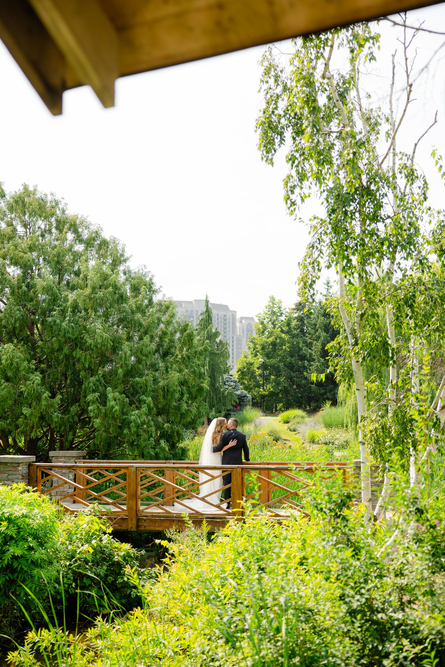 Distant view of wedding couple on wooden bridge surrounded by summer garden foliage.