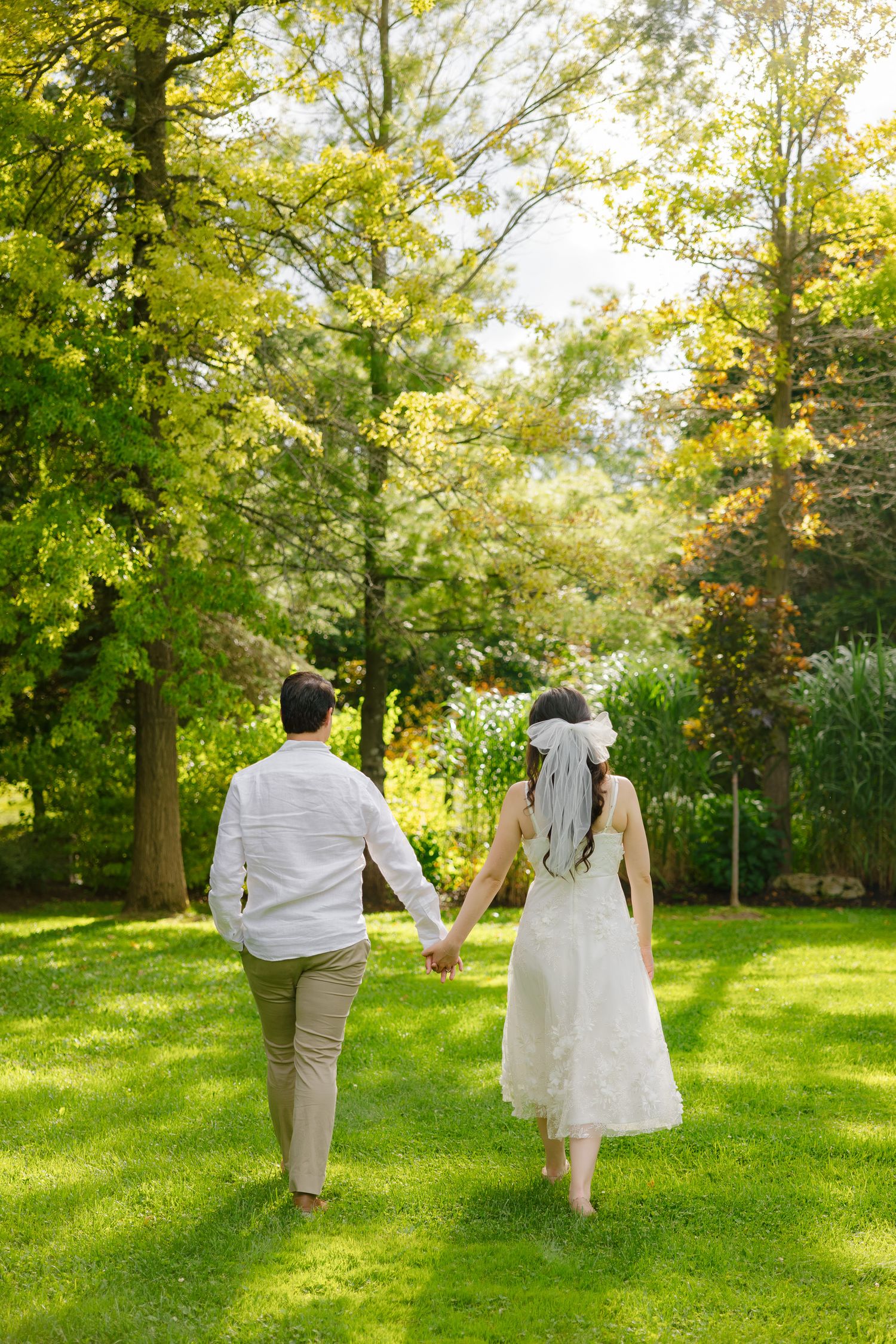 Newlyweds walking together on a green lawn surrounded by tall trees in golden sunlight.