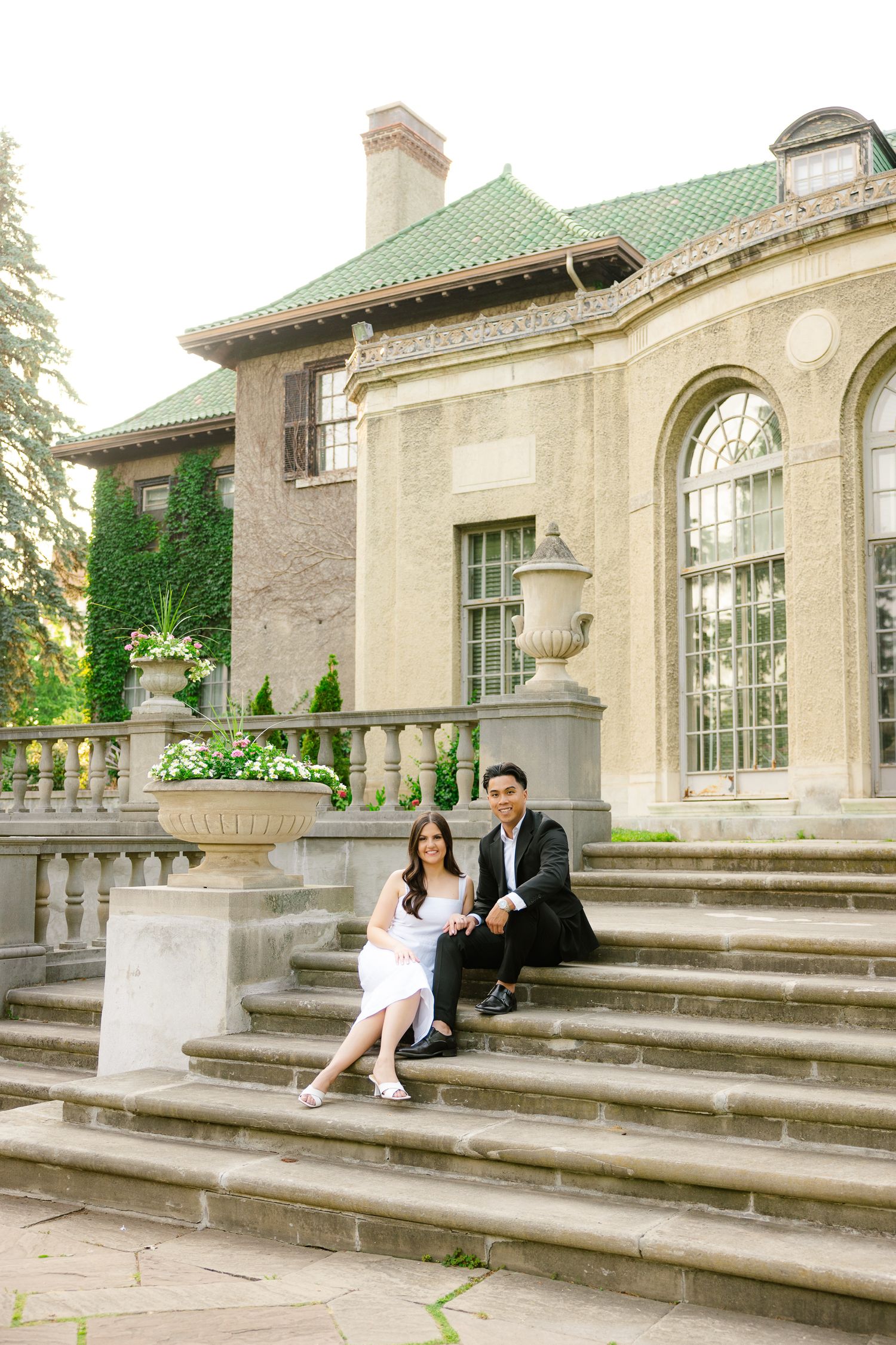Couple poses on grand stone steps of historic building with green copper roof and arched windows.