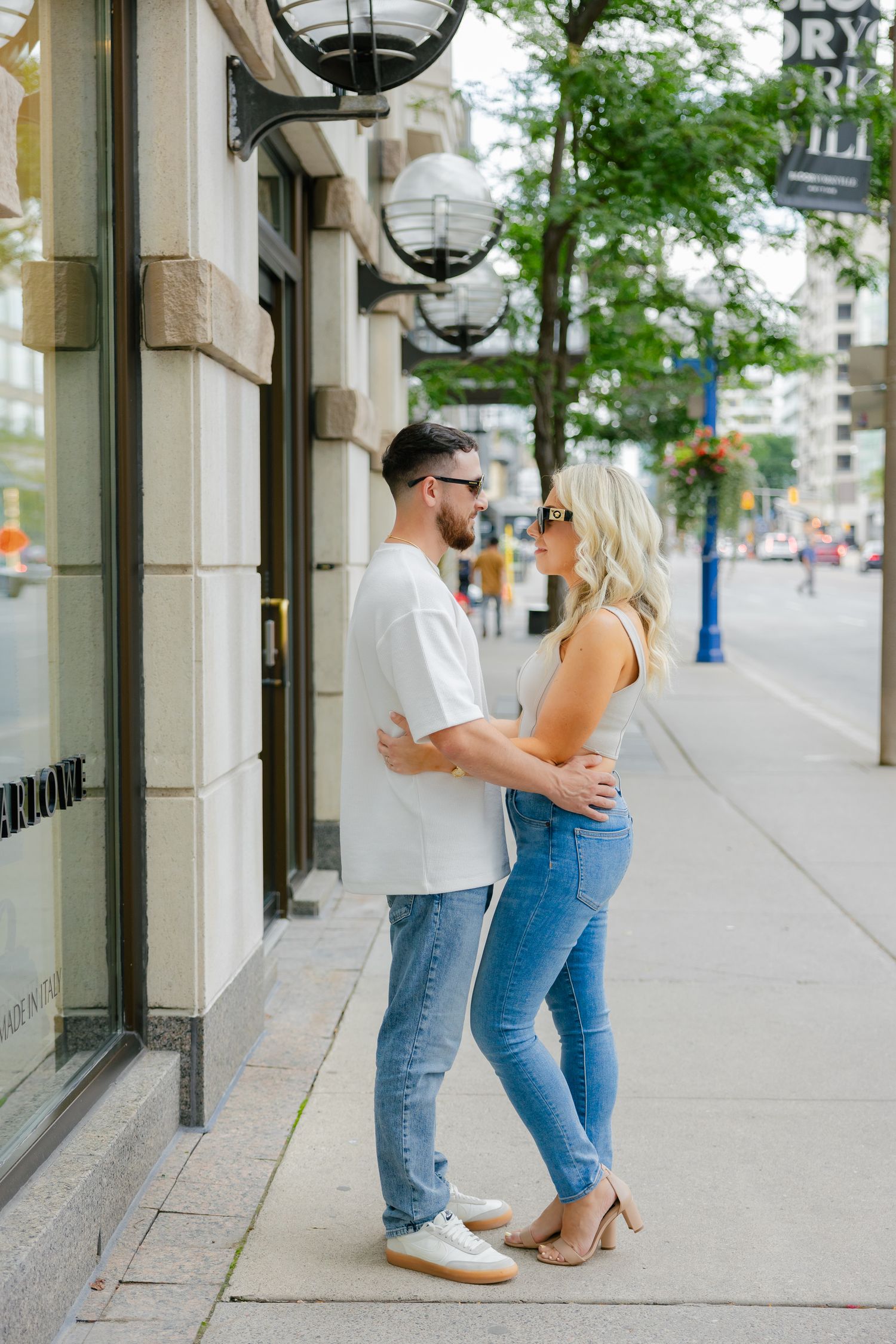 A romantic couple embraces on a city sidewalk near decorative street lamps.