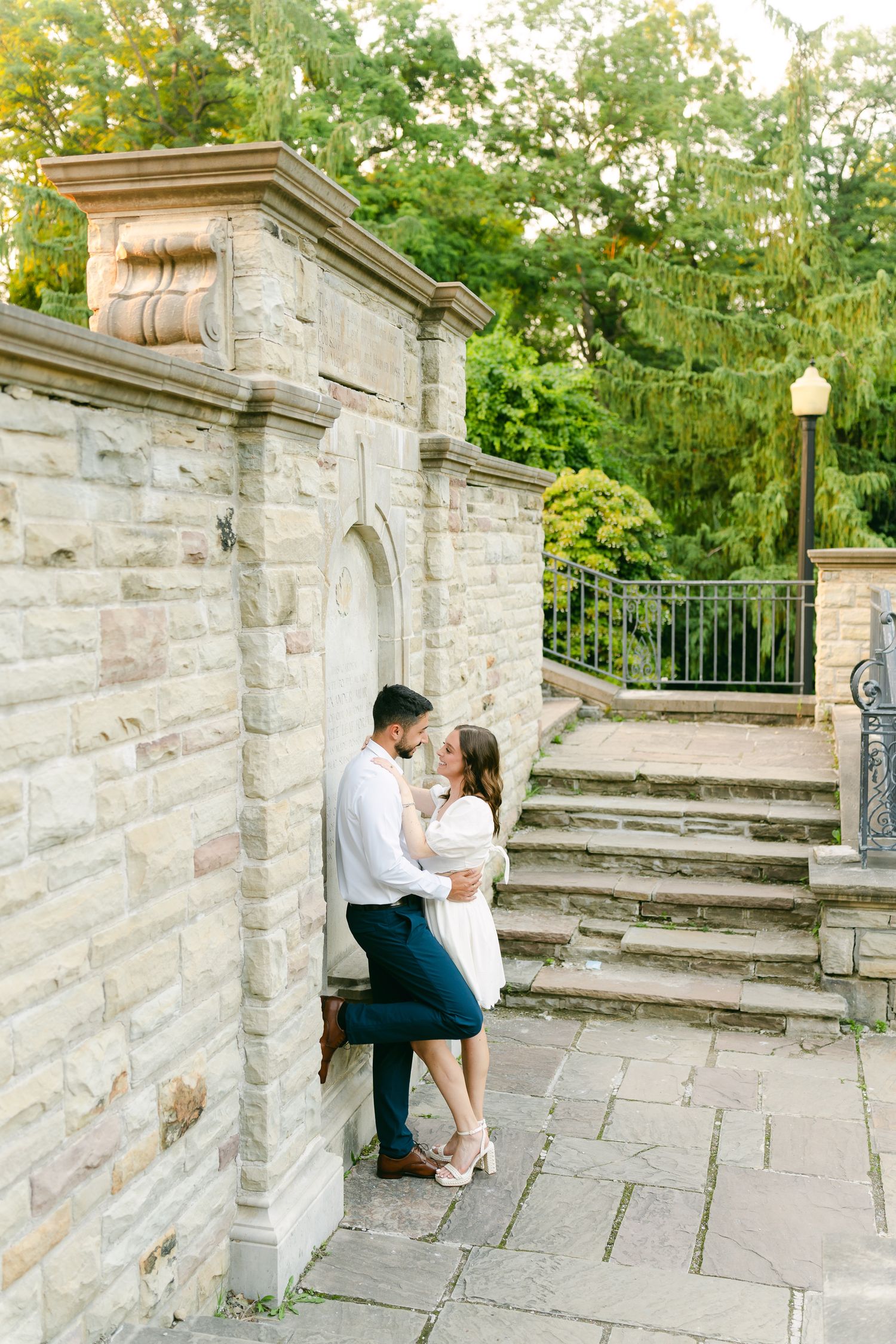 A couple embraces against a stone wall near garden steps on a sunny day.
