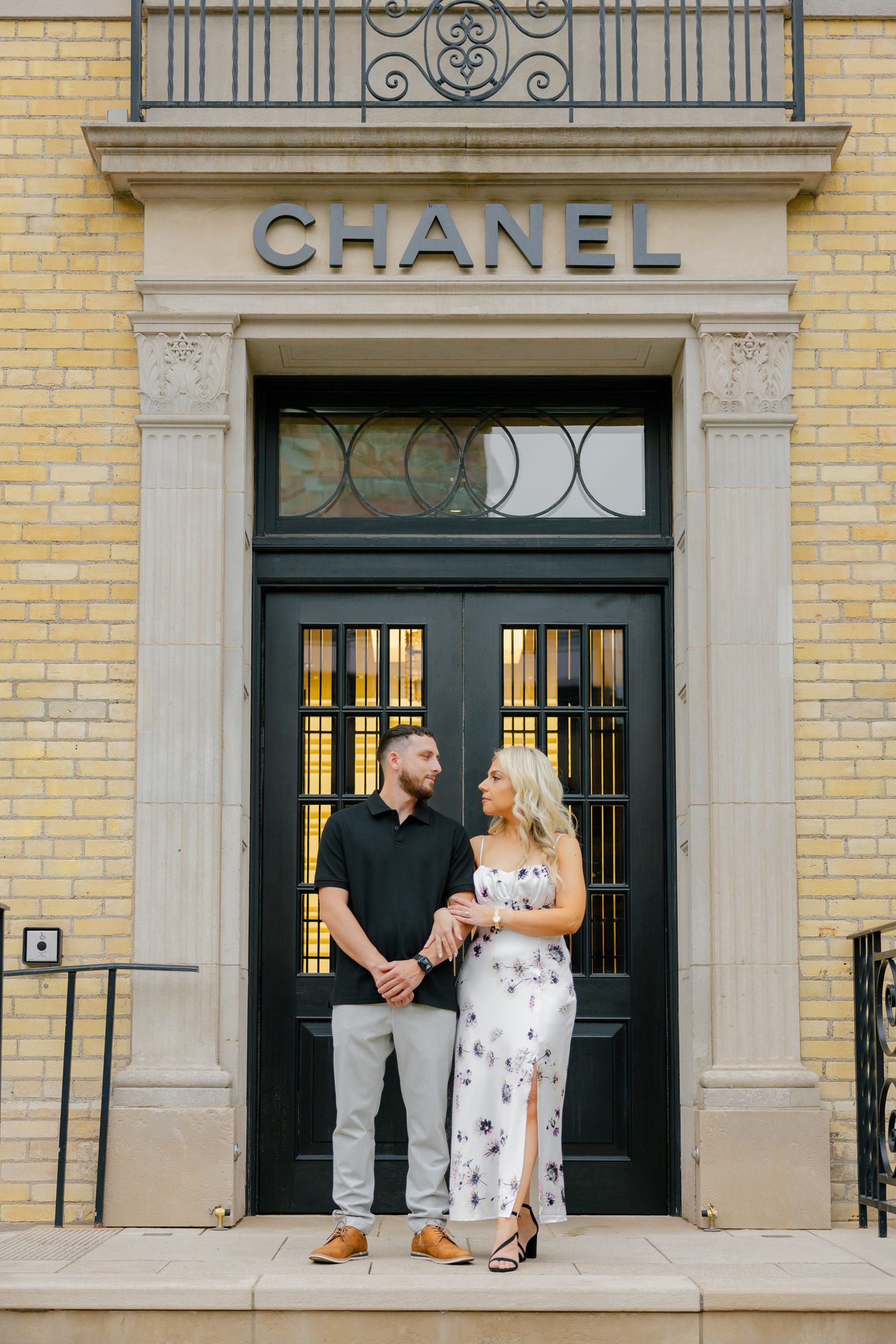 Couple poses elegantly at the entrance of a Chanel store with yellow brick facade.
