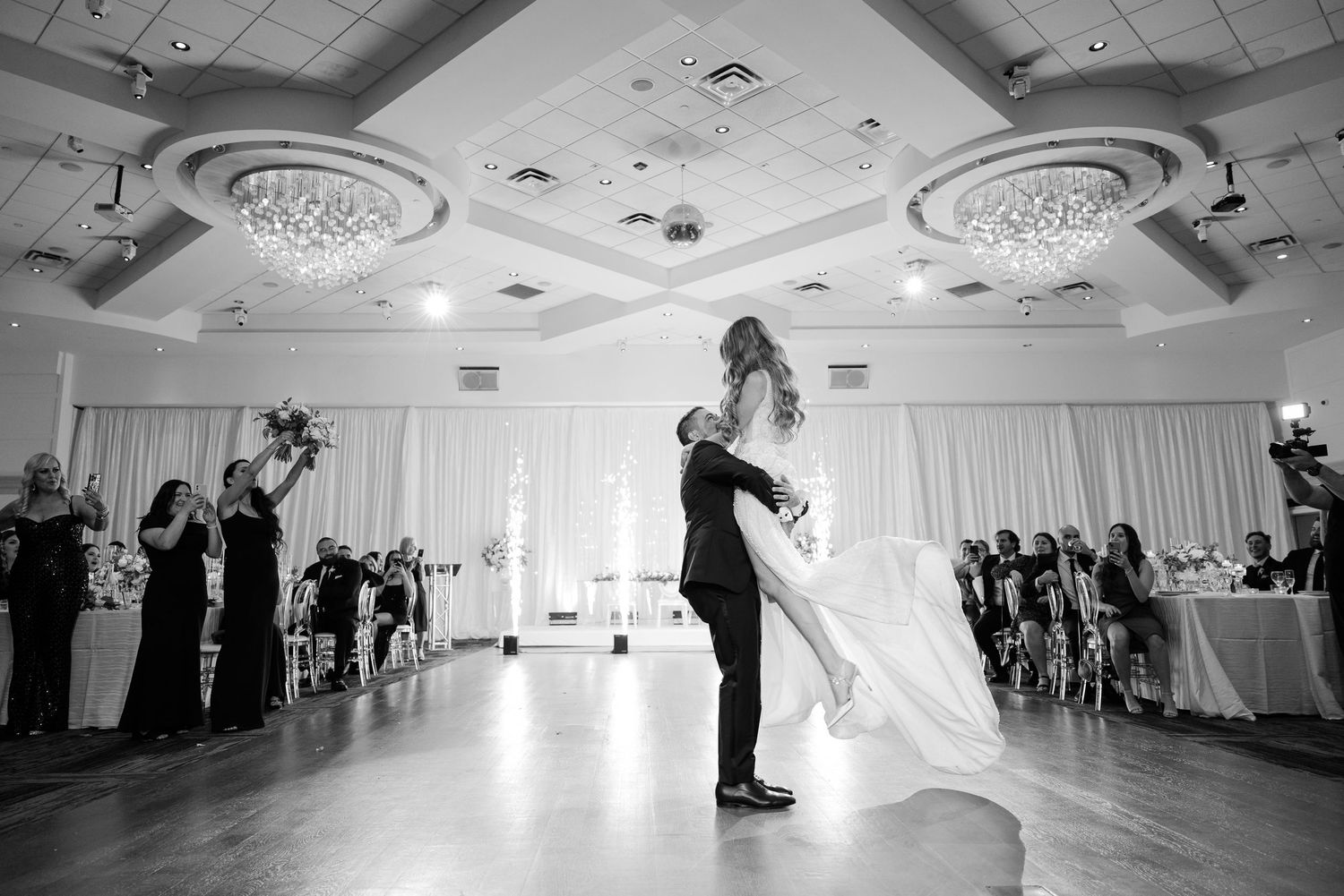 Black and white photo of romantic first dance in elegant ballroom with ornate ceiling details.