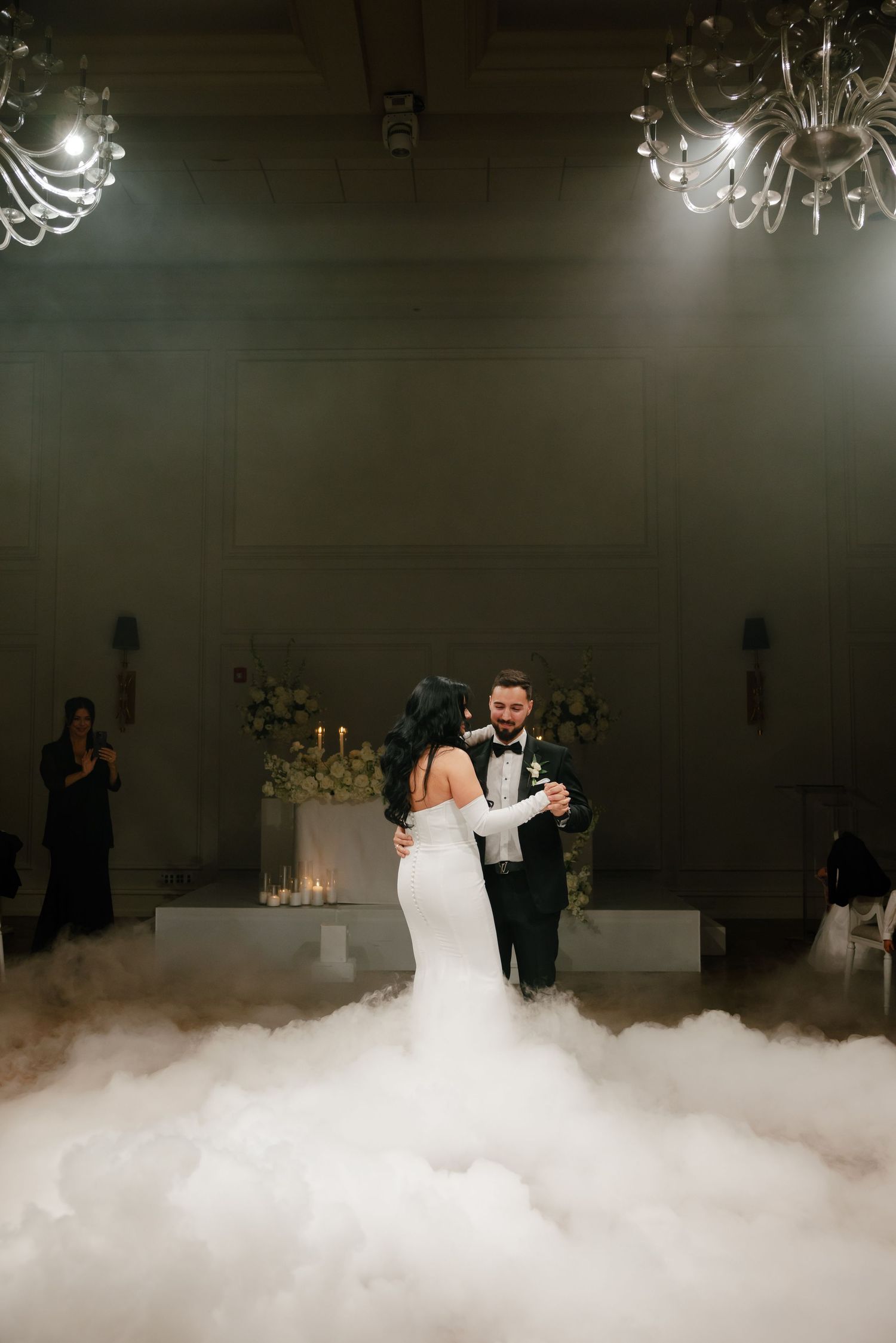 Couple shares their first dance surrounded by atmospheric fog effects and elegant chandeliers at their wedding reception.