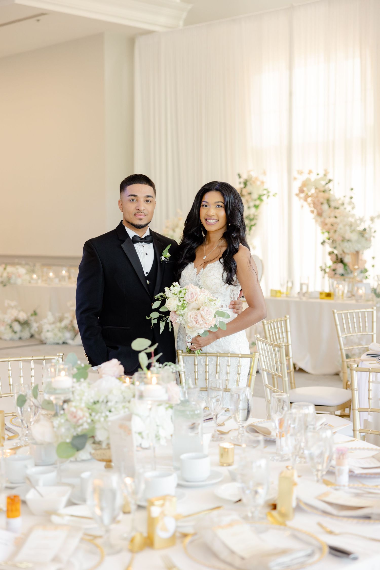 Wedding couple posing by their reception table with white floral centerpieces and gold chiavari chairs.