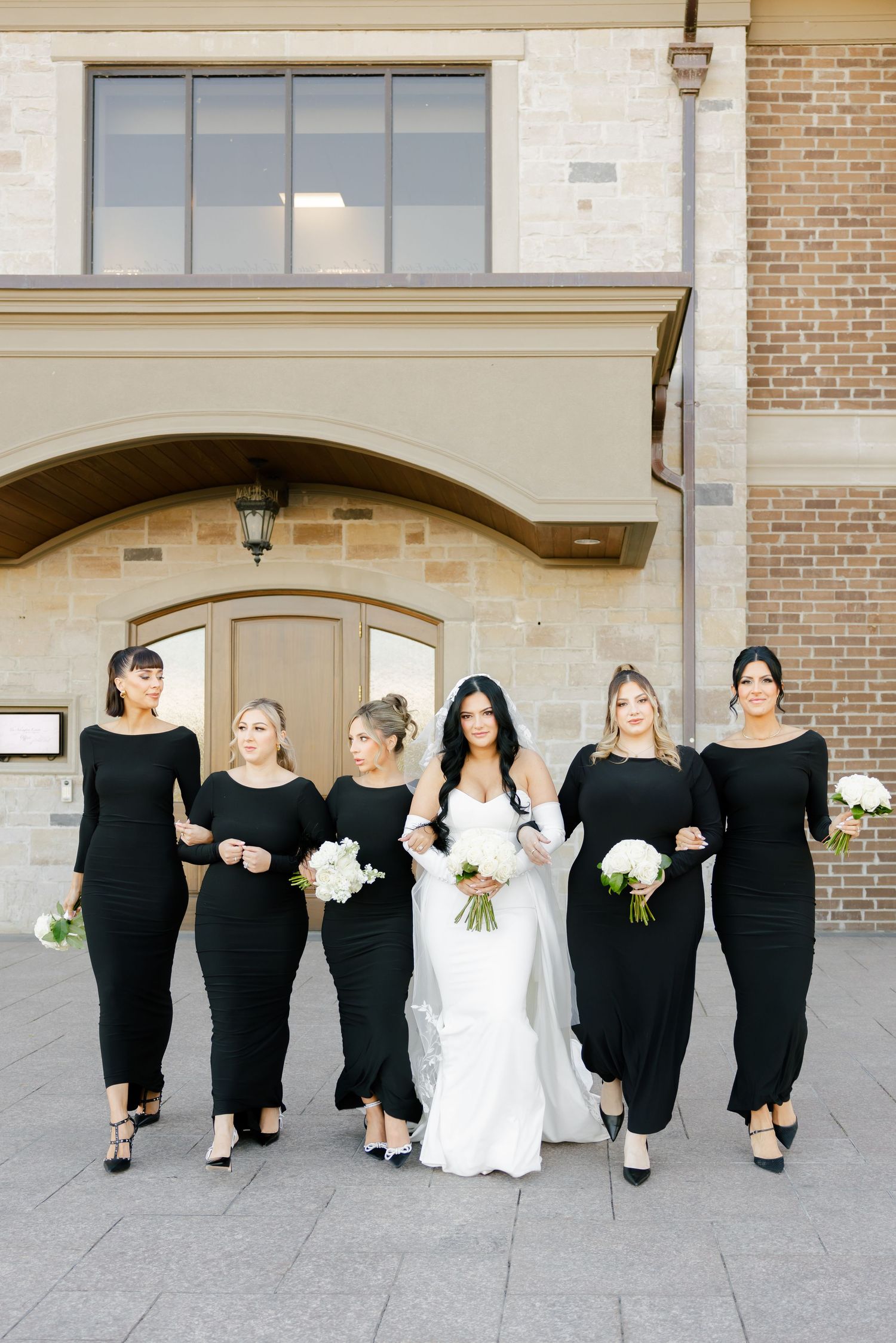 Modern bridal party in black gowns poses with bride in front of stone building.