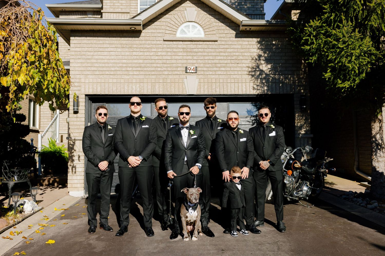 A group wearing formal black attire poses with a dog in front of a beige house.