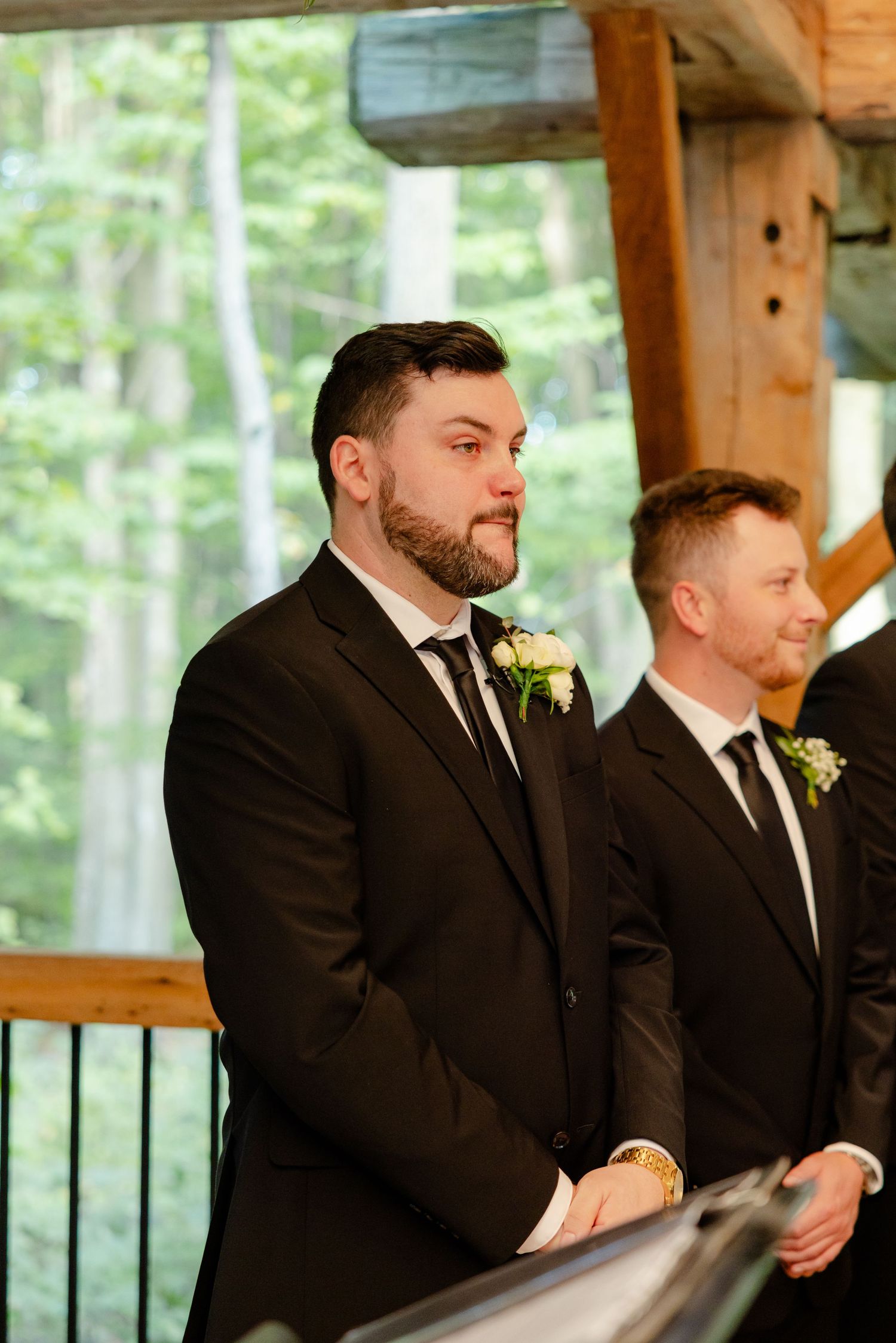 Groomsmen in black suits with boutonnières stand at attention during outdoor forest wedding ceremony.