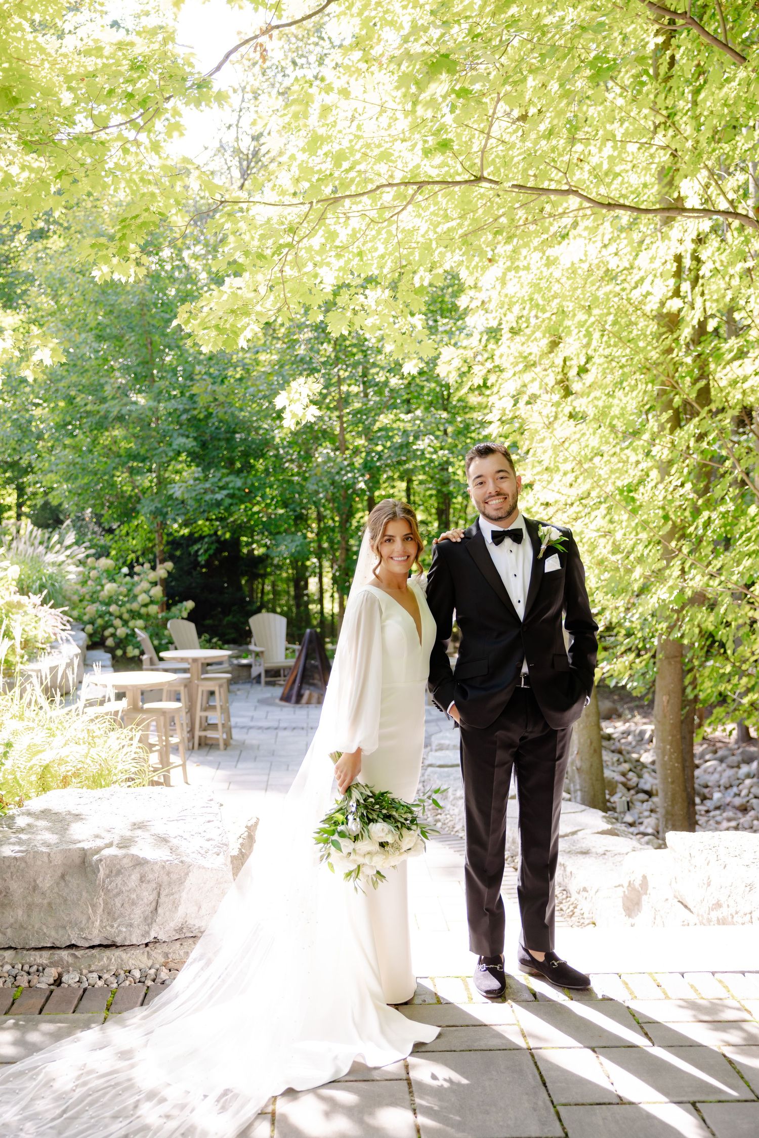 Elegant bride in white gown and groom in tuxedo pose together on stone patio surrounded by lush greenery.