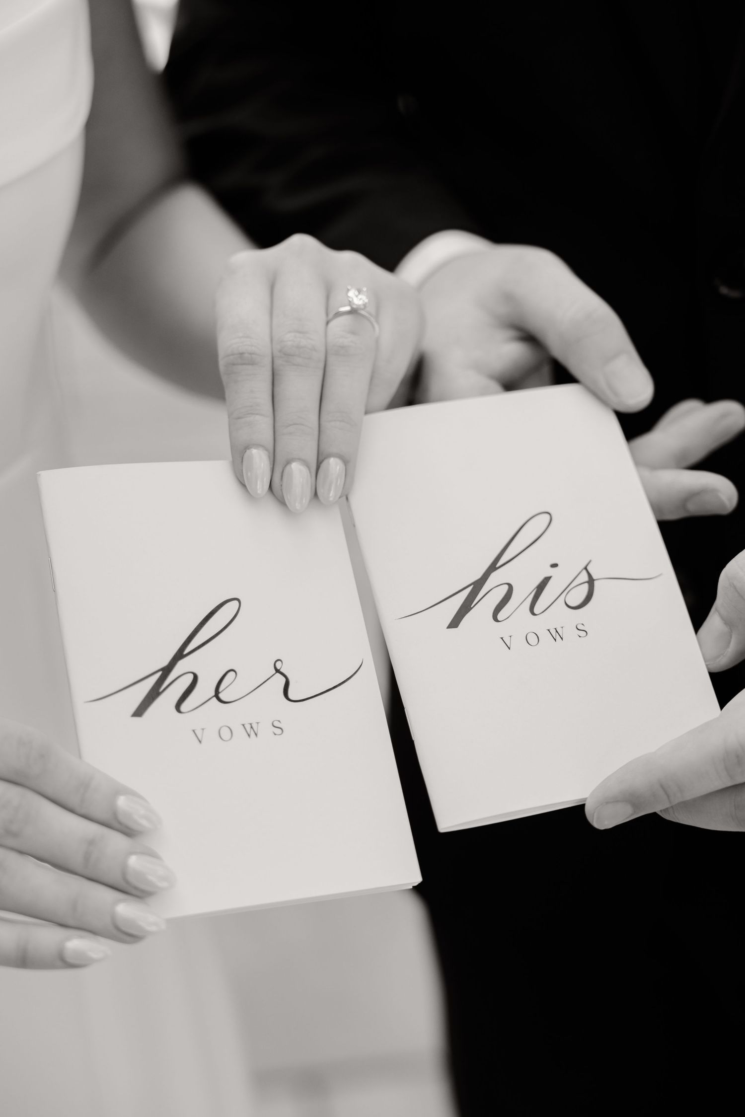 Black and white detail shot of wedding vow books labeled 'his' and 'her' with hands showing engagement ring.