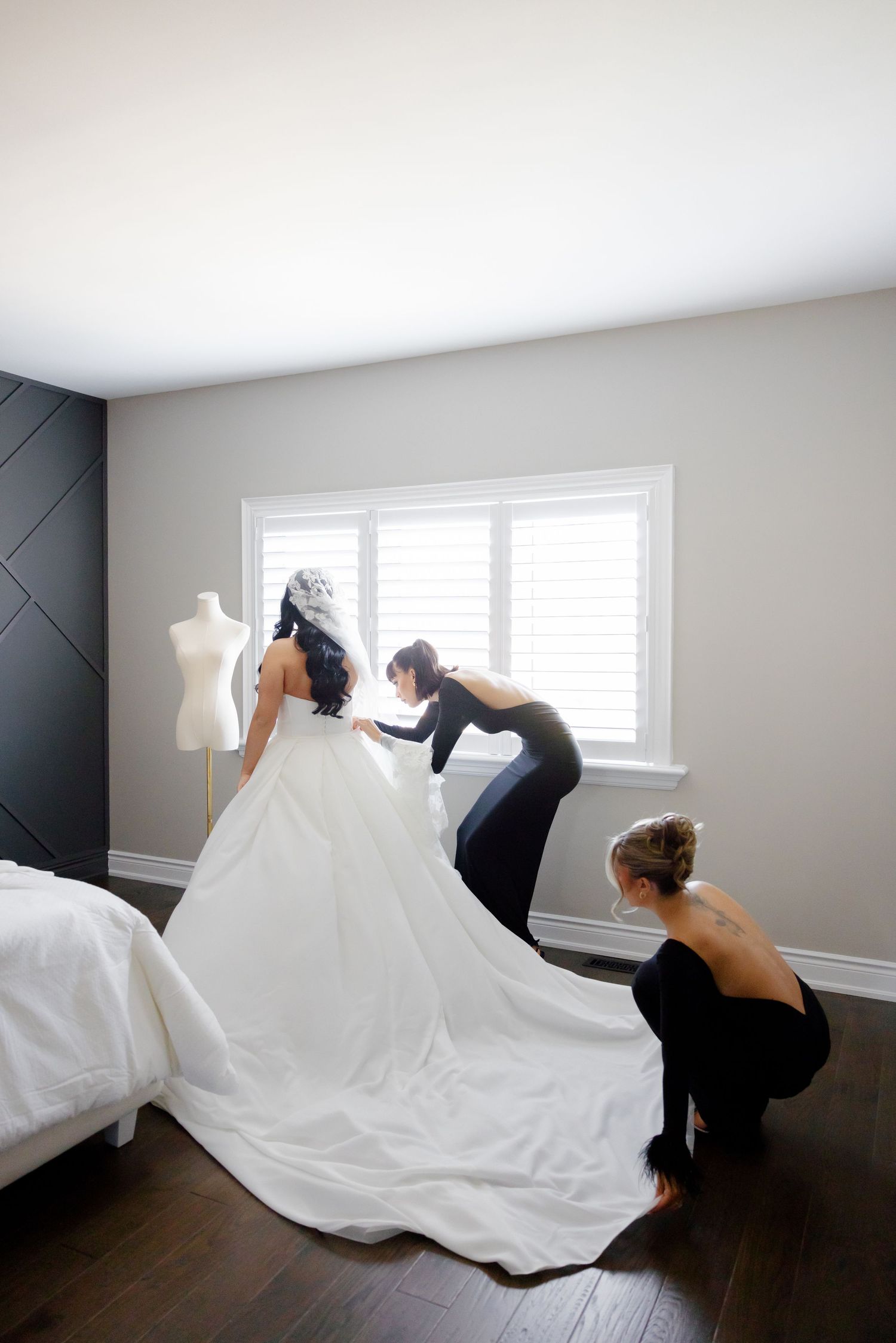 Bridesmaids help adjust the long train of an elegant white wedding dress in a modern bedroom.