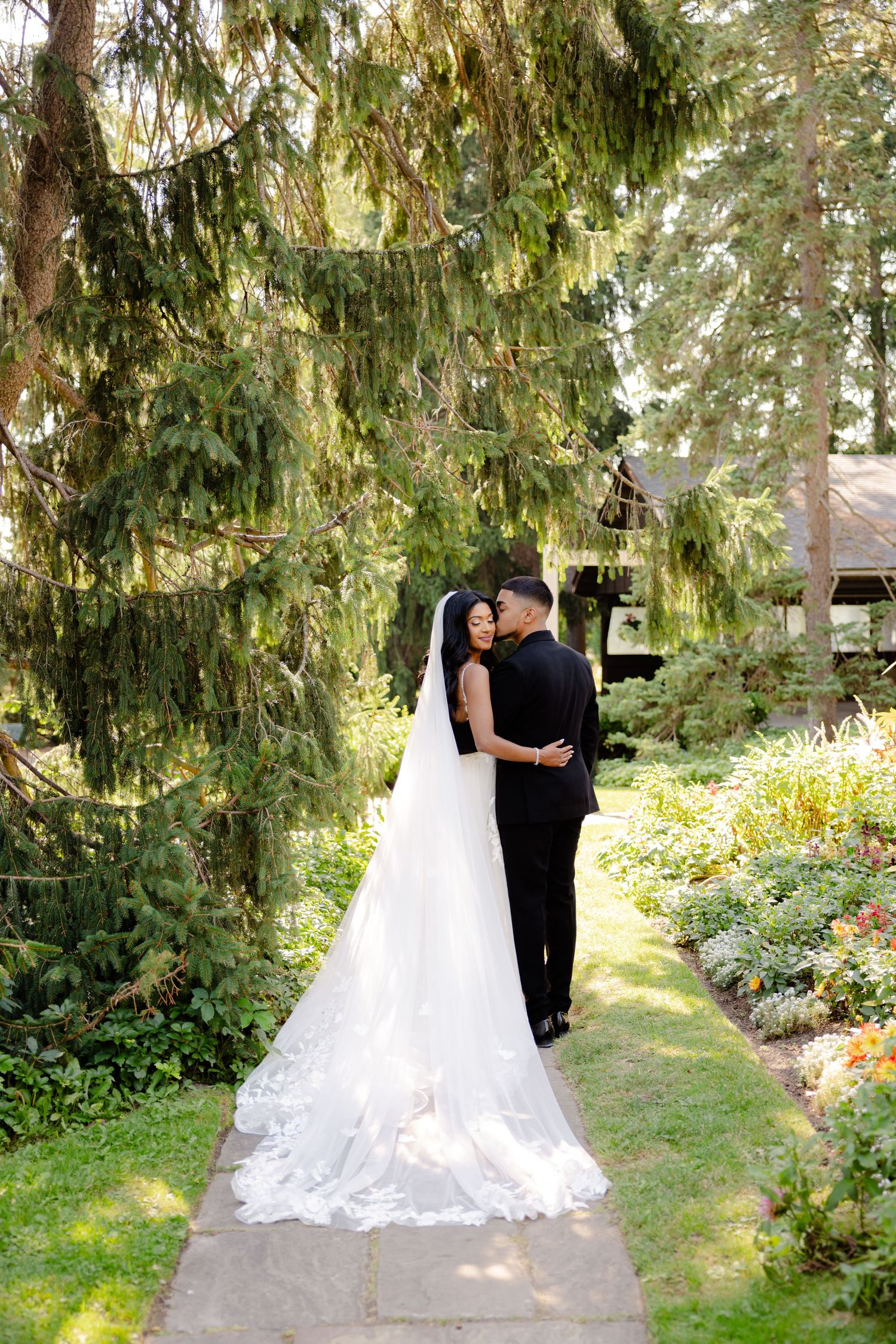 A couple embraces on a garden pathway beneath dramatic drooping evergreen branches.