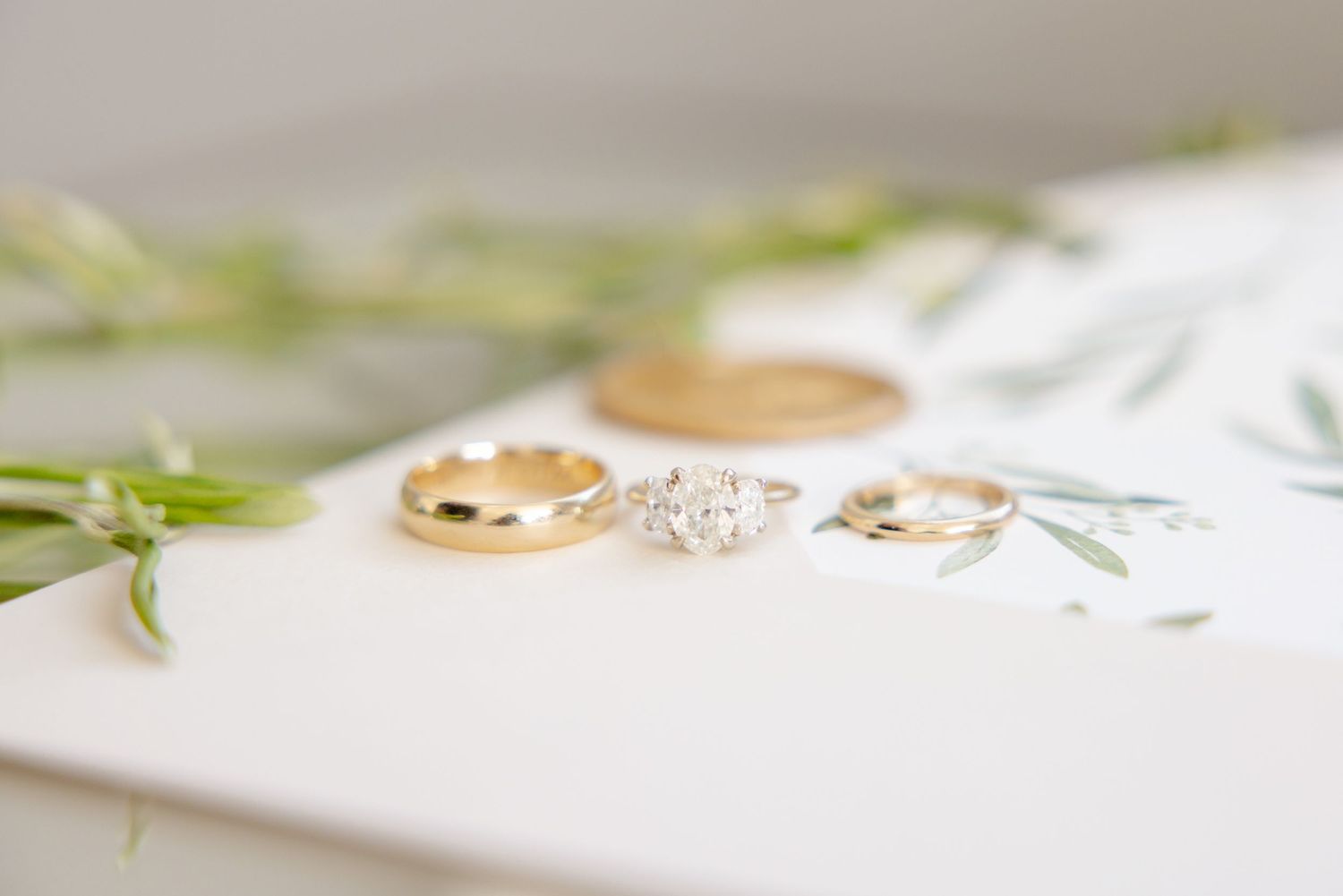 Three gold and diamond wedding rings arranged with delicate greenery on white surface.