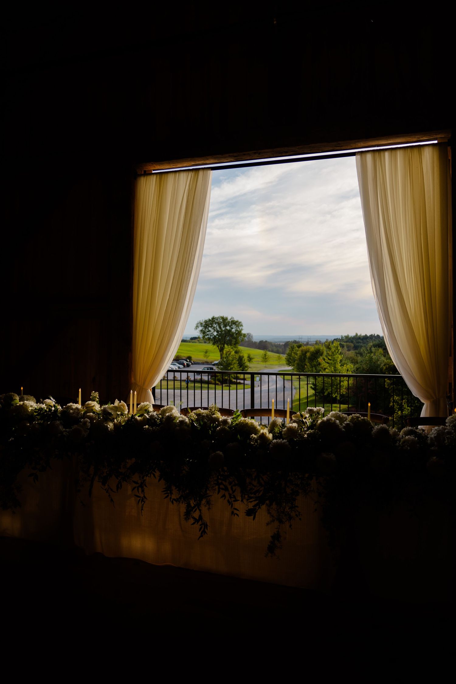 A dramatic window view of rolling hills framed by cream curtains and floral decorations.