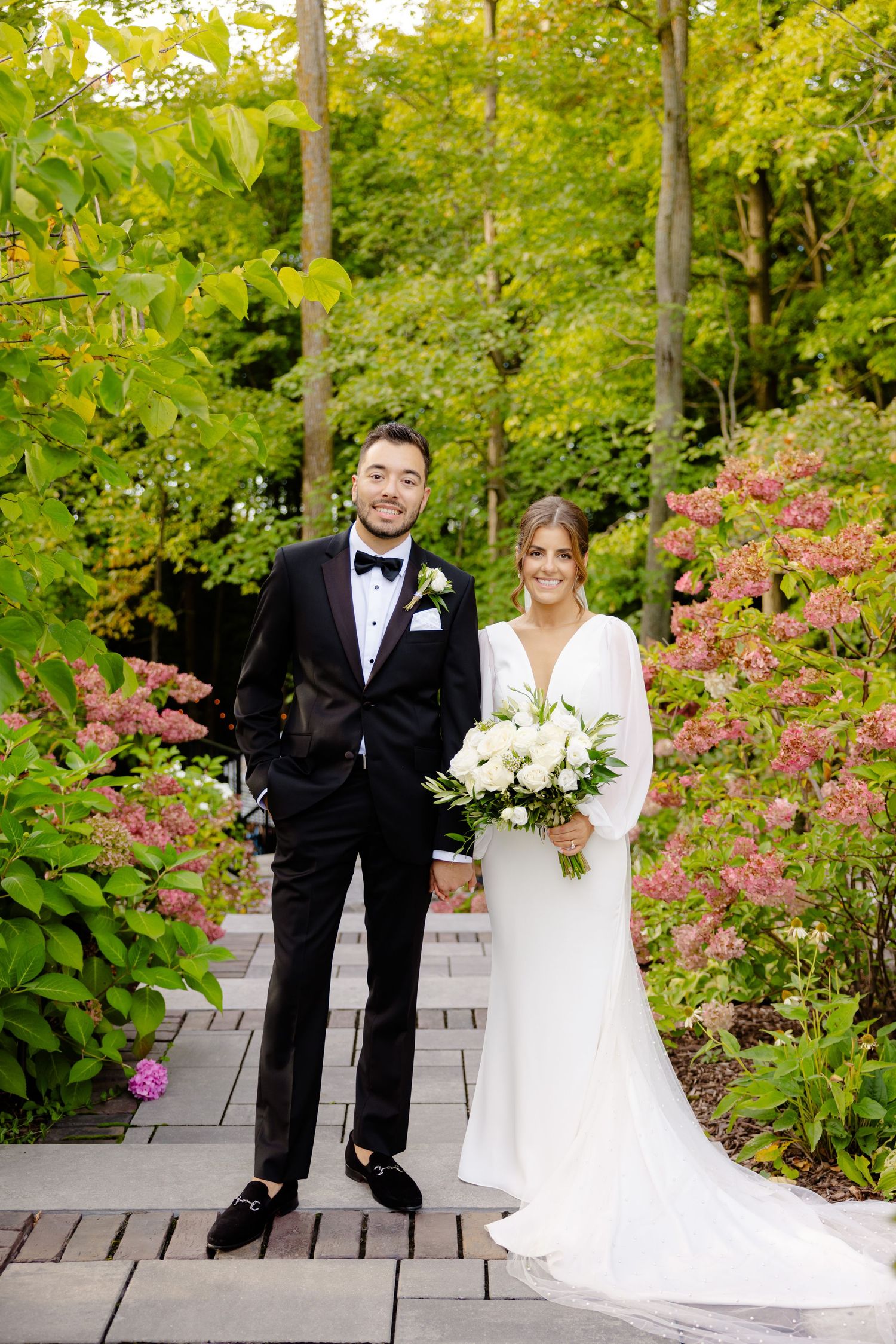 Elegant wedding portrait on wooden pathway with pink azaleas and green foliage backdrop.