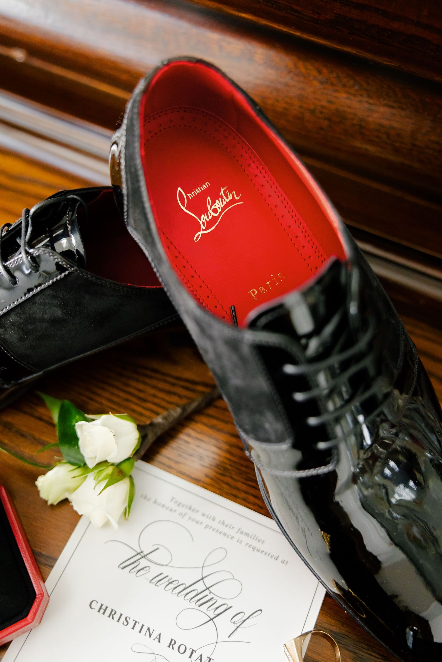 Black patent leather dress shoe with red interior lining next to elegant wedding invitation on wooden surface.