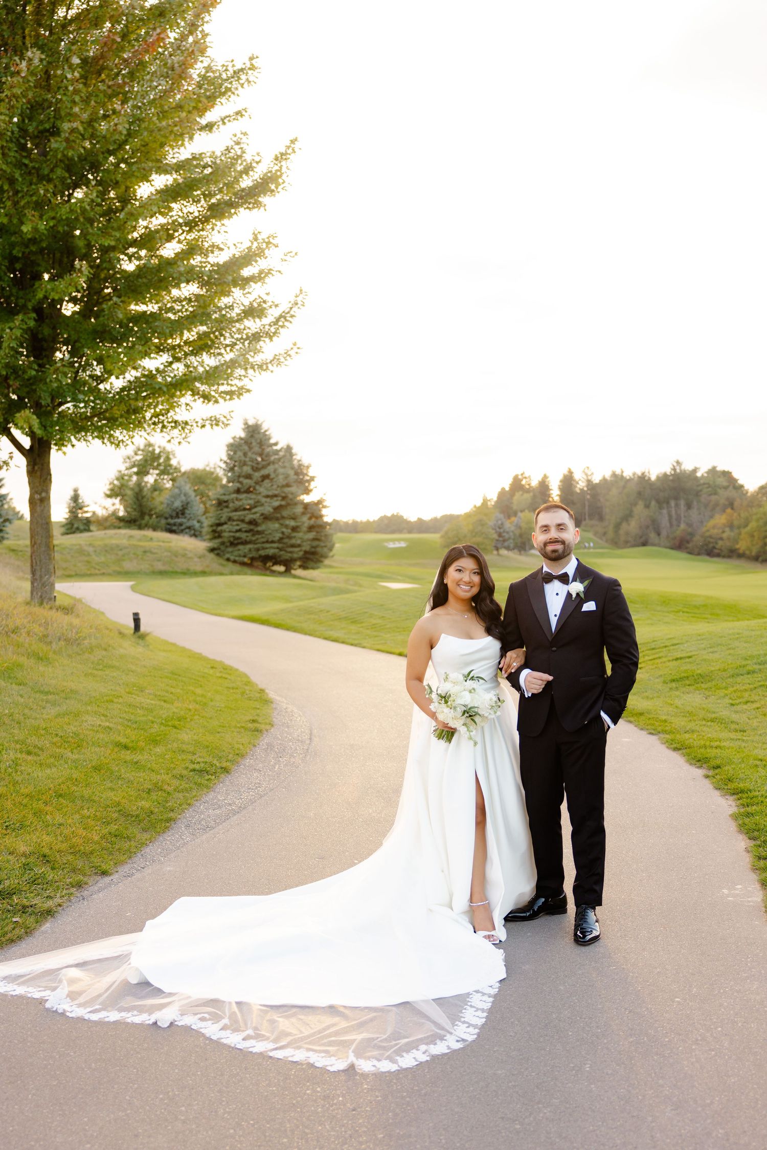 Newlyweds walk together on a winding golf course path with a flowing cathedral veil trailing behind.