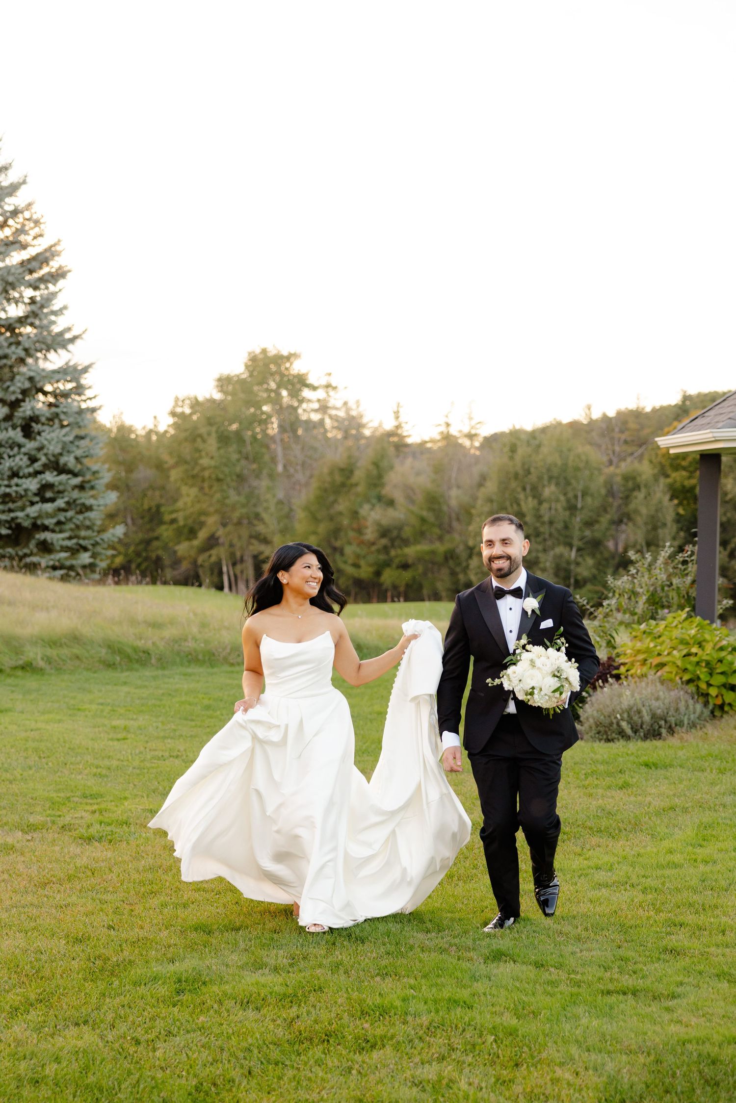 Wedding couple strolling across manicured lawn with flowing white dress and blue spruce trees in background.
