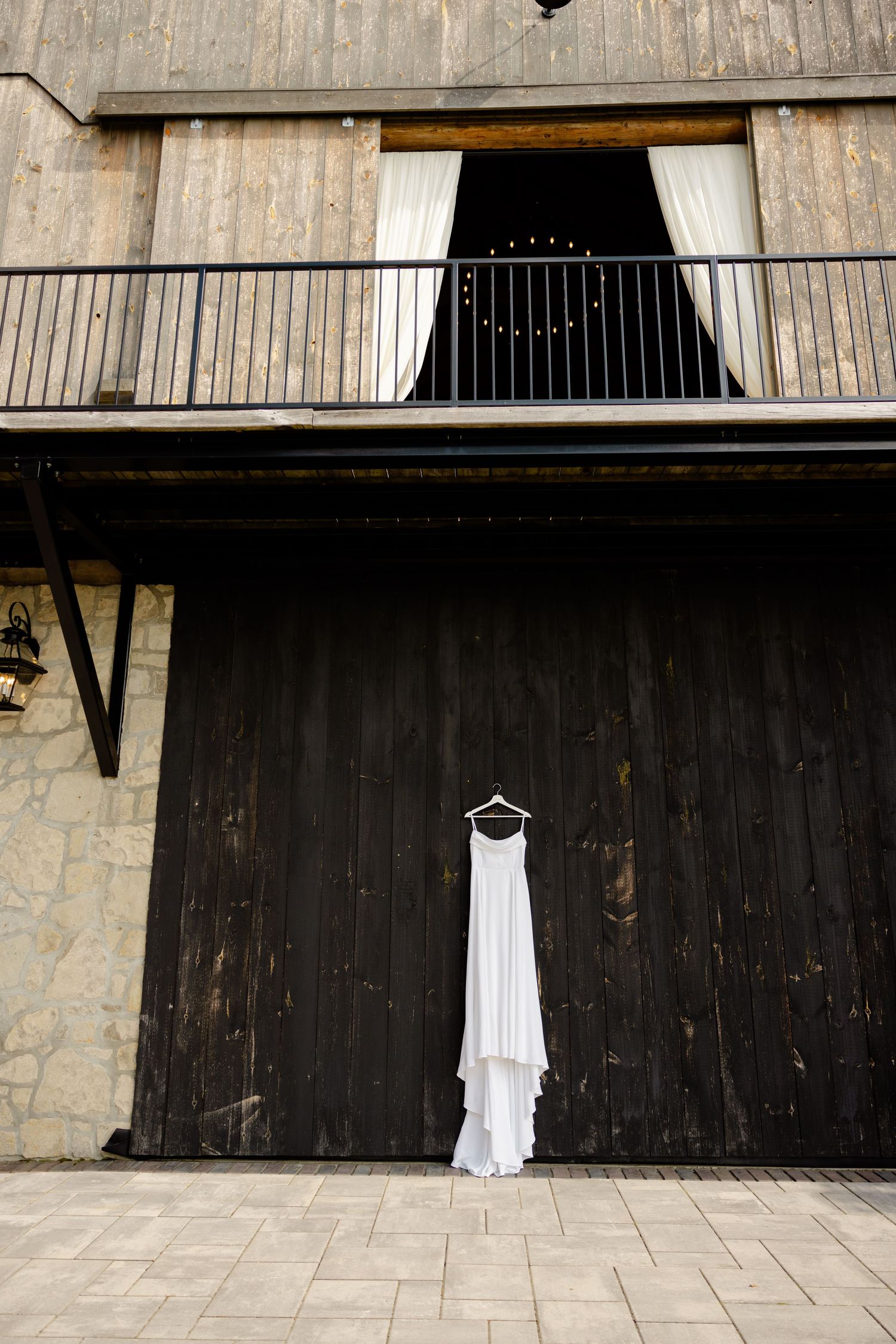 Wedding dress displayed against rustic barn wall with balcony and white curtains.