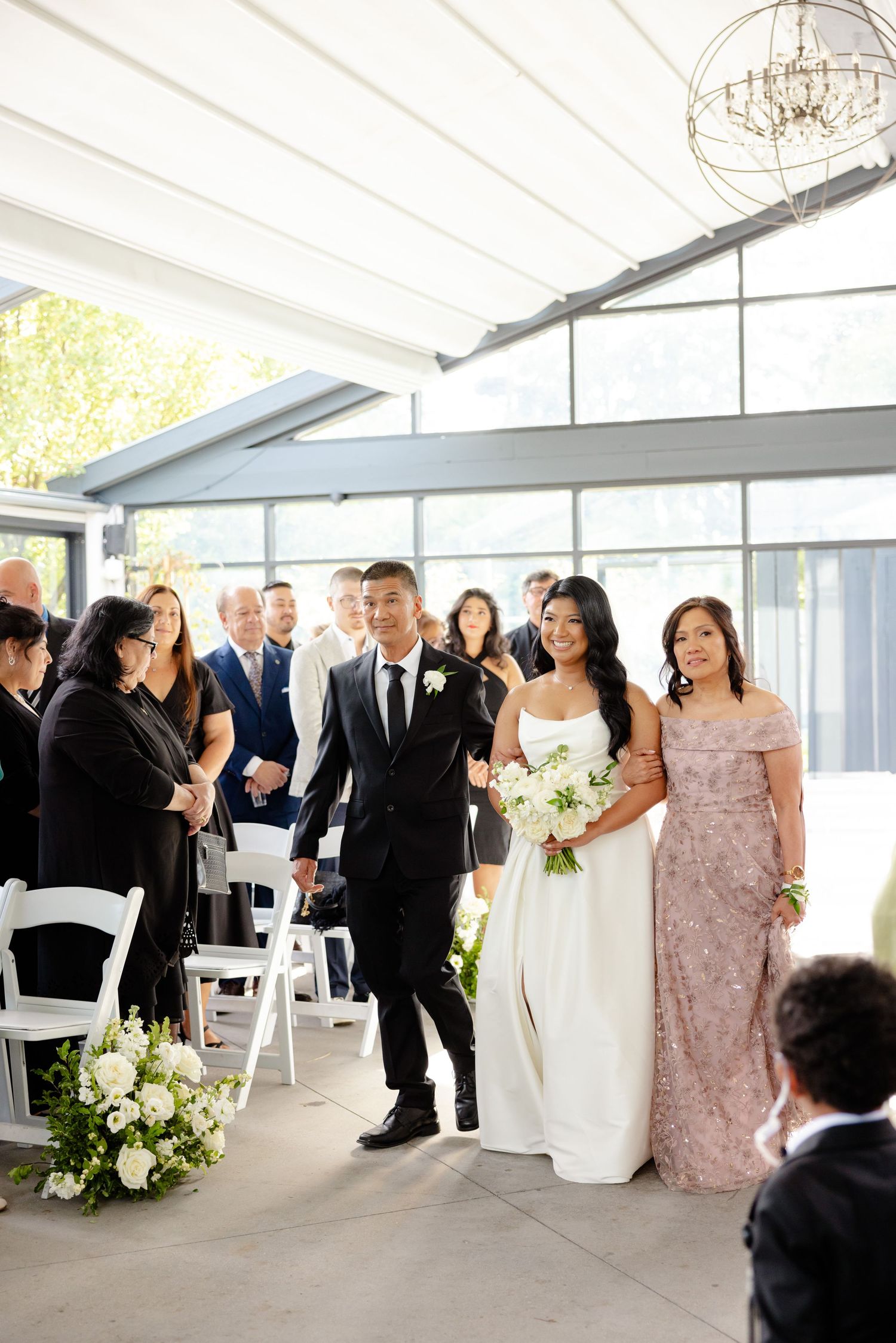 Guests look on as a wedding party processes through a modern indoor venue with large windows.