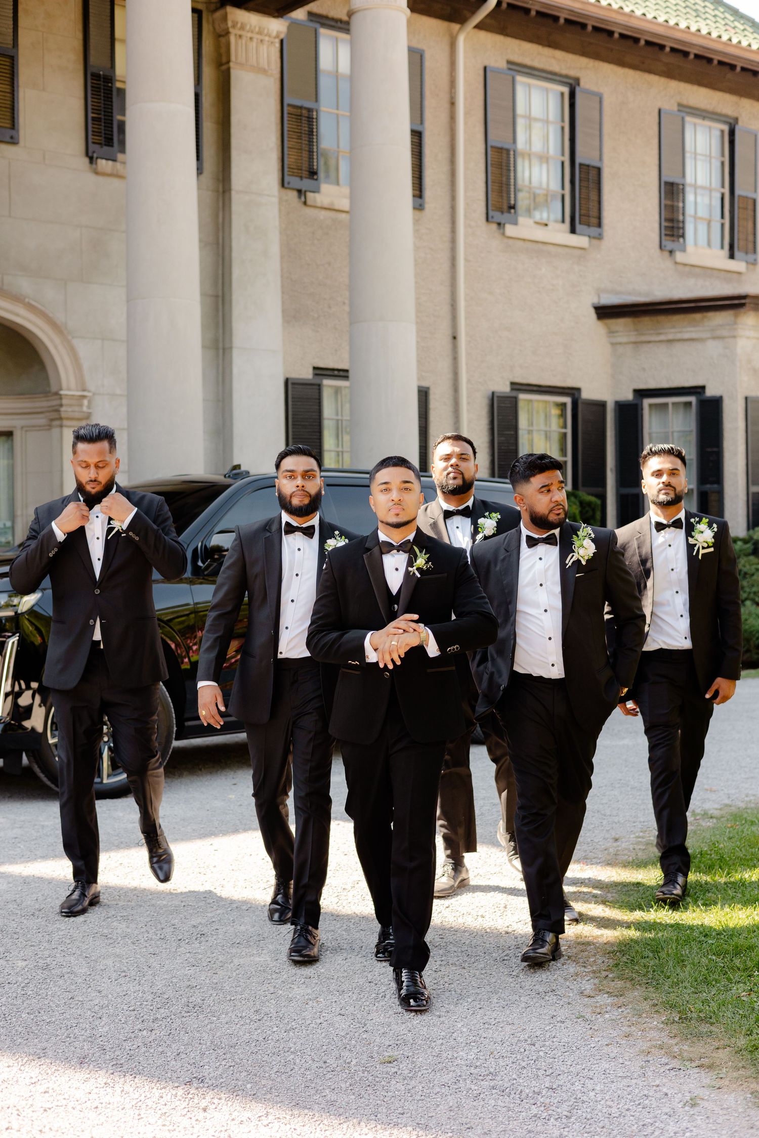 Stylish groomsmen in black tuxedos walking together outside a grand building.