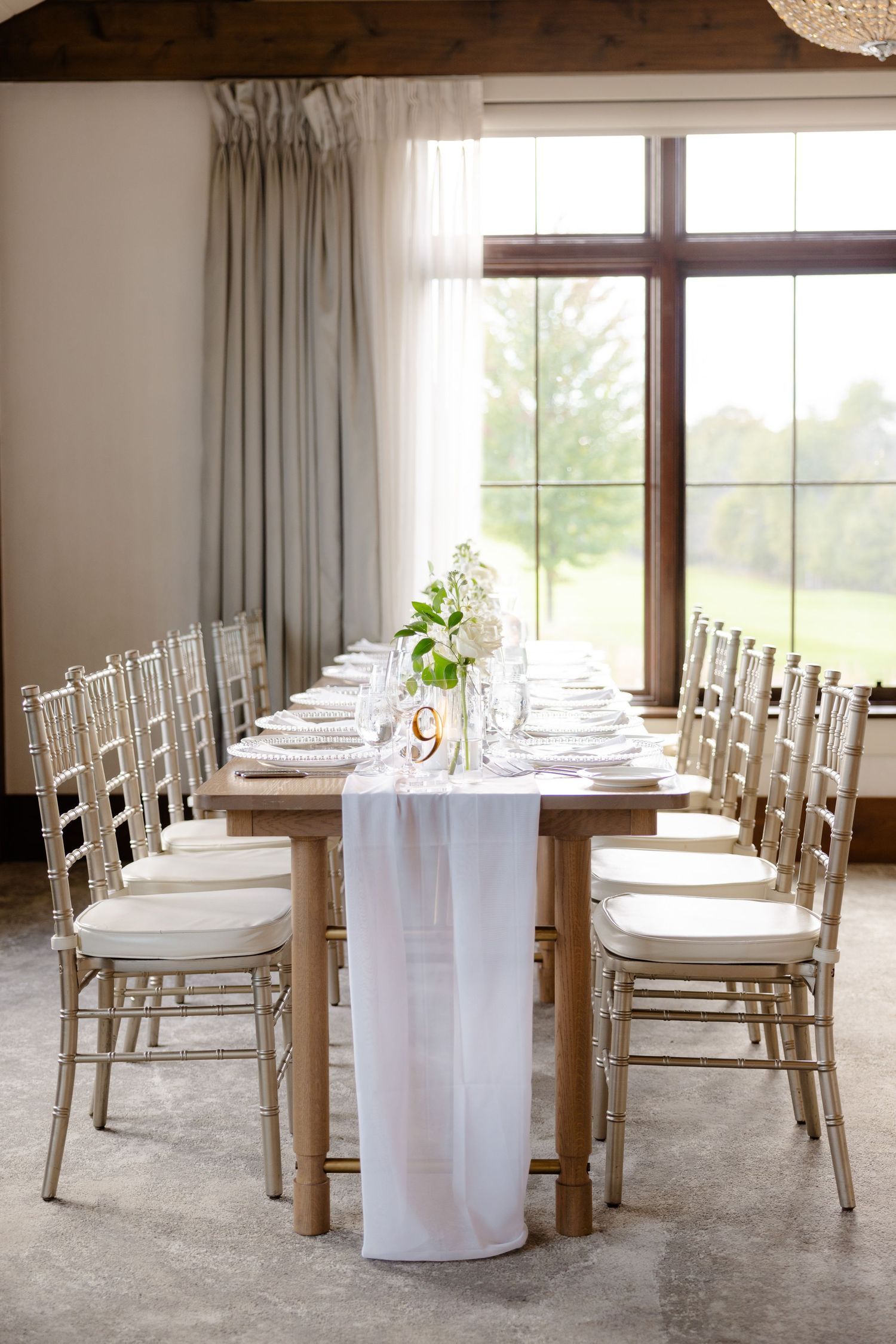 Long dining table with white runner and gold chiavari chairs set for wedding reception.