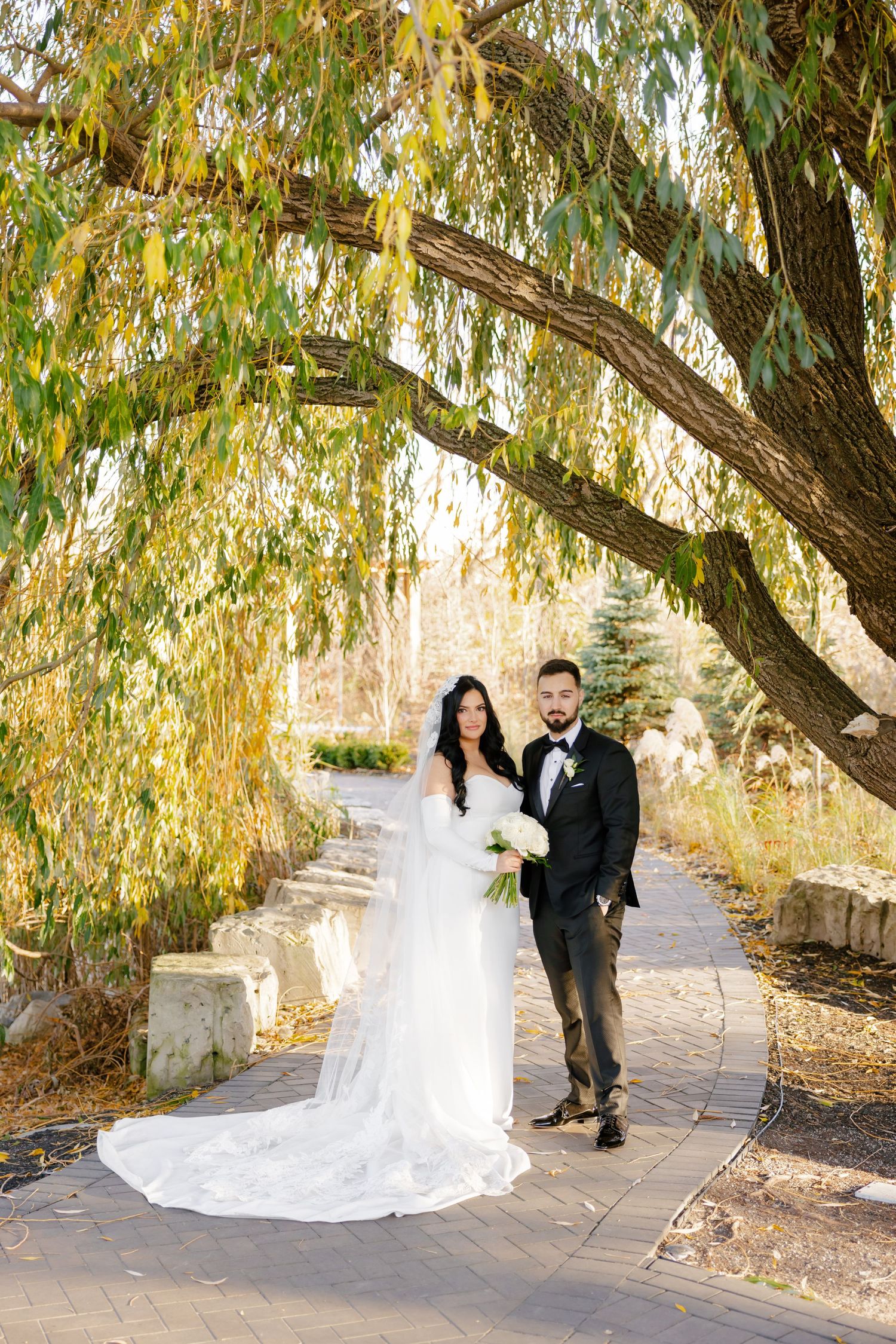 A bride in white gown and groom in tuxedo pose under autumn willow tree branches.